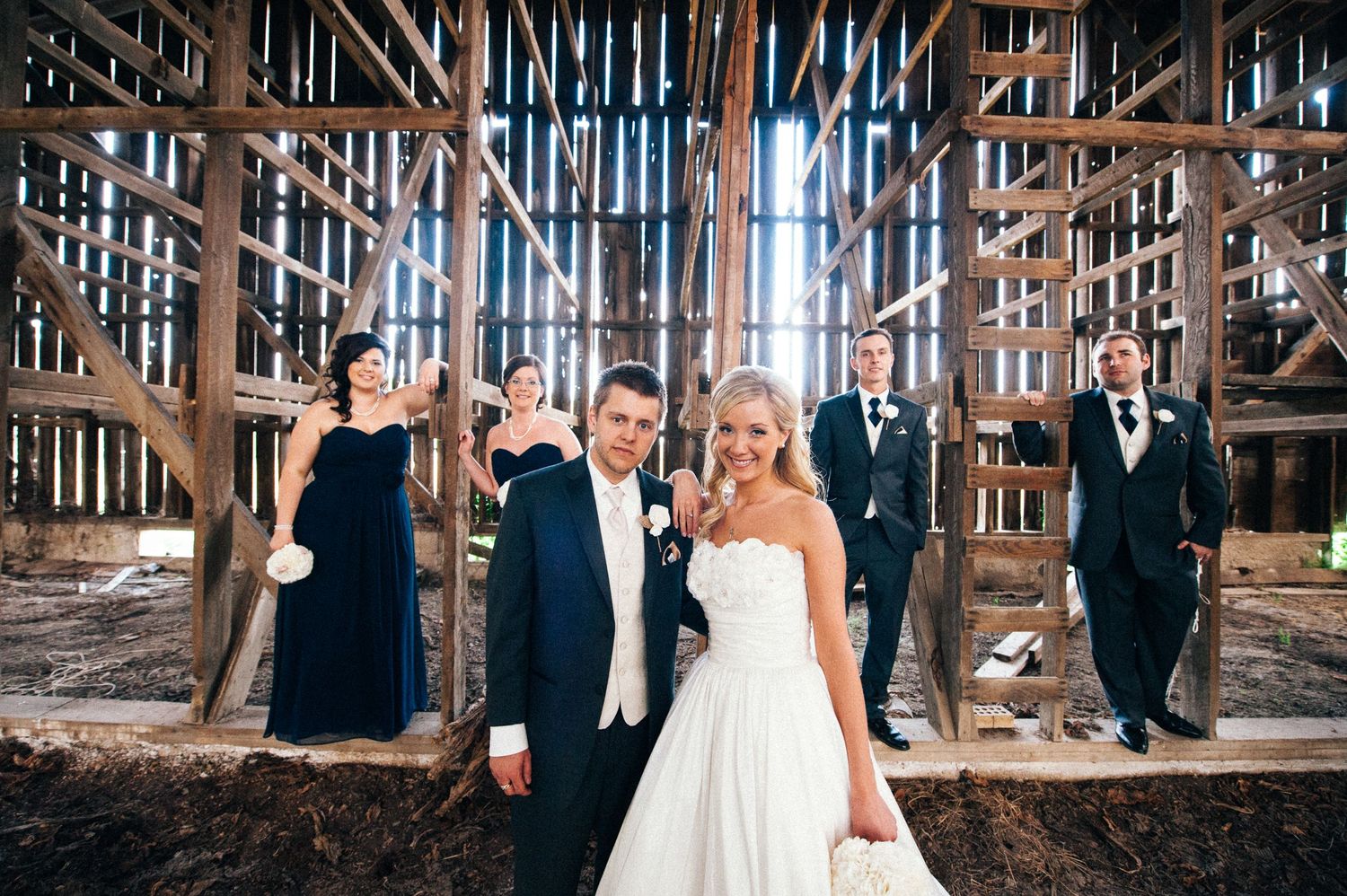 Wedding party poses in front of rustic wooden barn structure in formal attire.
