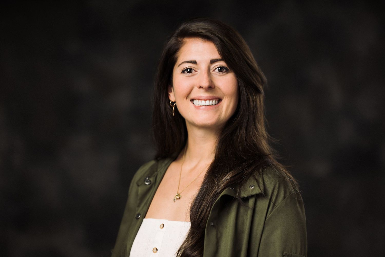 Professional headshot of someone in a dark olive green jacket against a dark background with warm lighting.