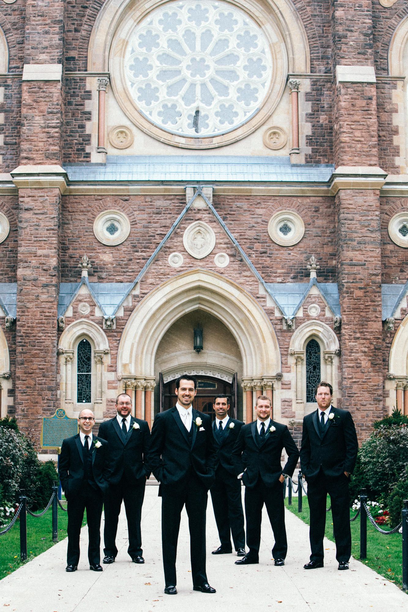 Wedding party in formal attire poses in front of a historic brick church with ornate rose window.