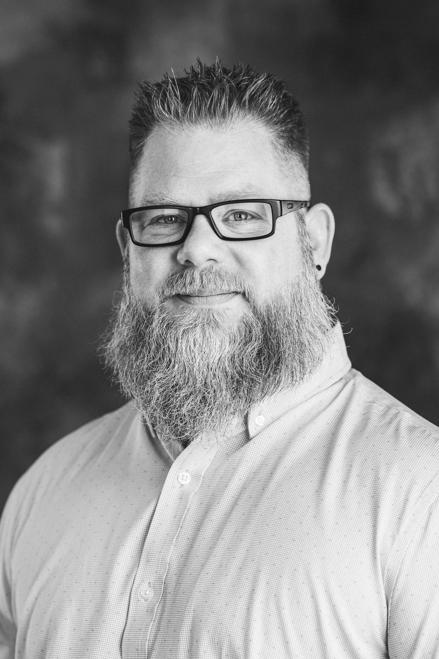 Man with beard and glasses in white shirt against dark background in professional headshots.