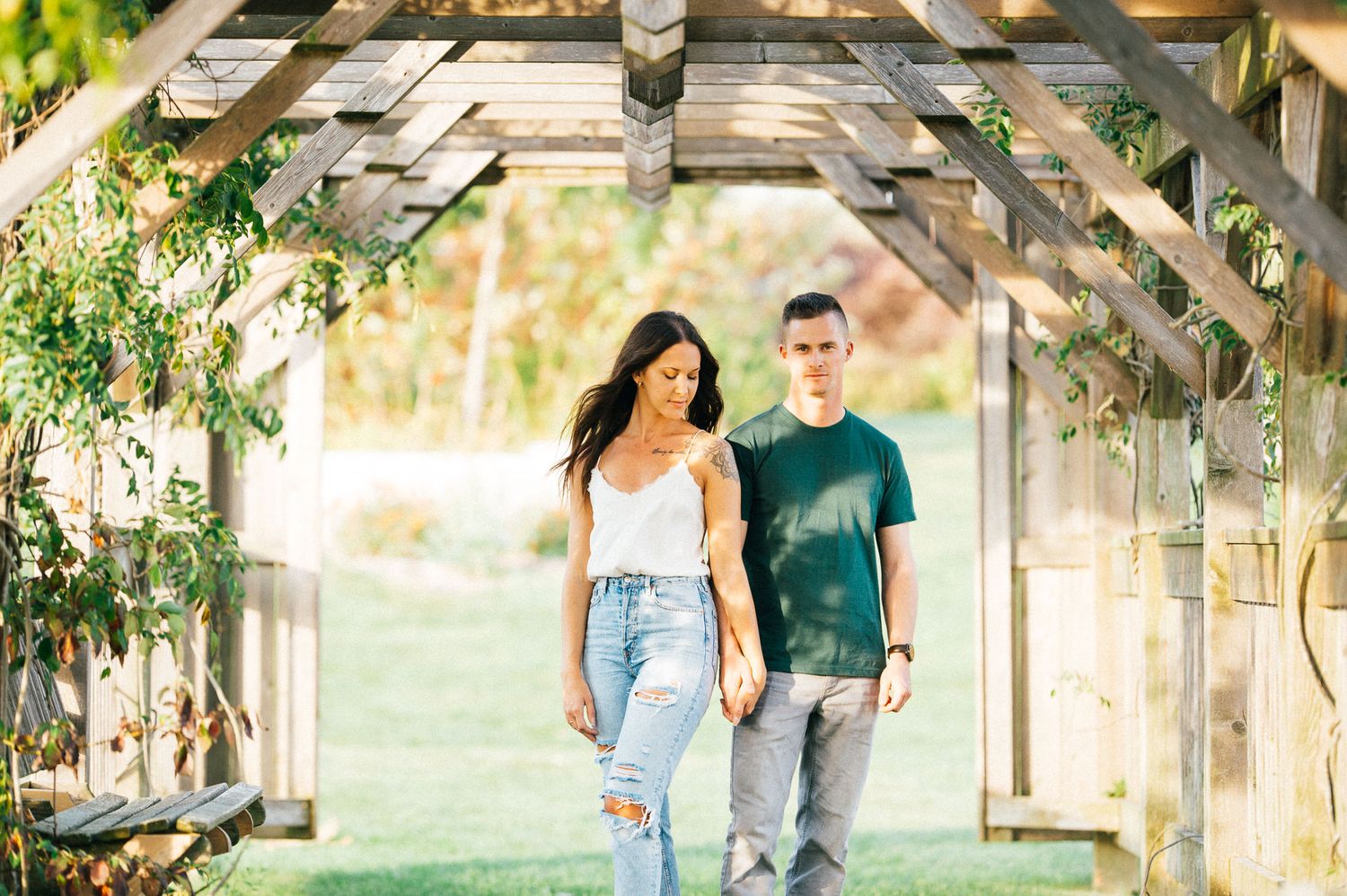 A couple walks together through a wooden pergola with hanging vines and natural lighting.