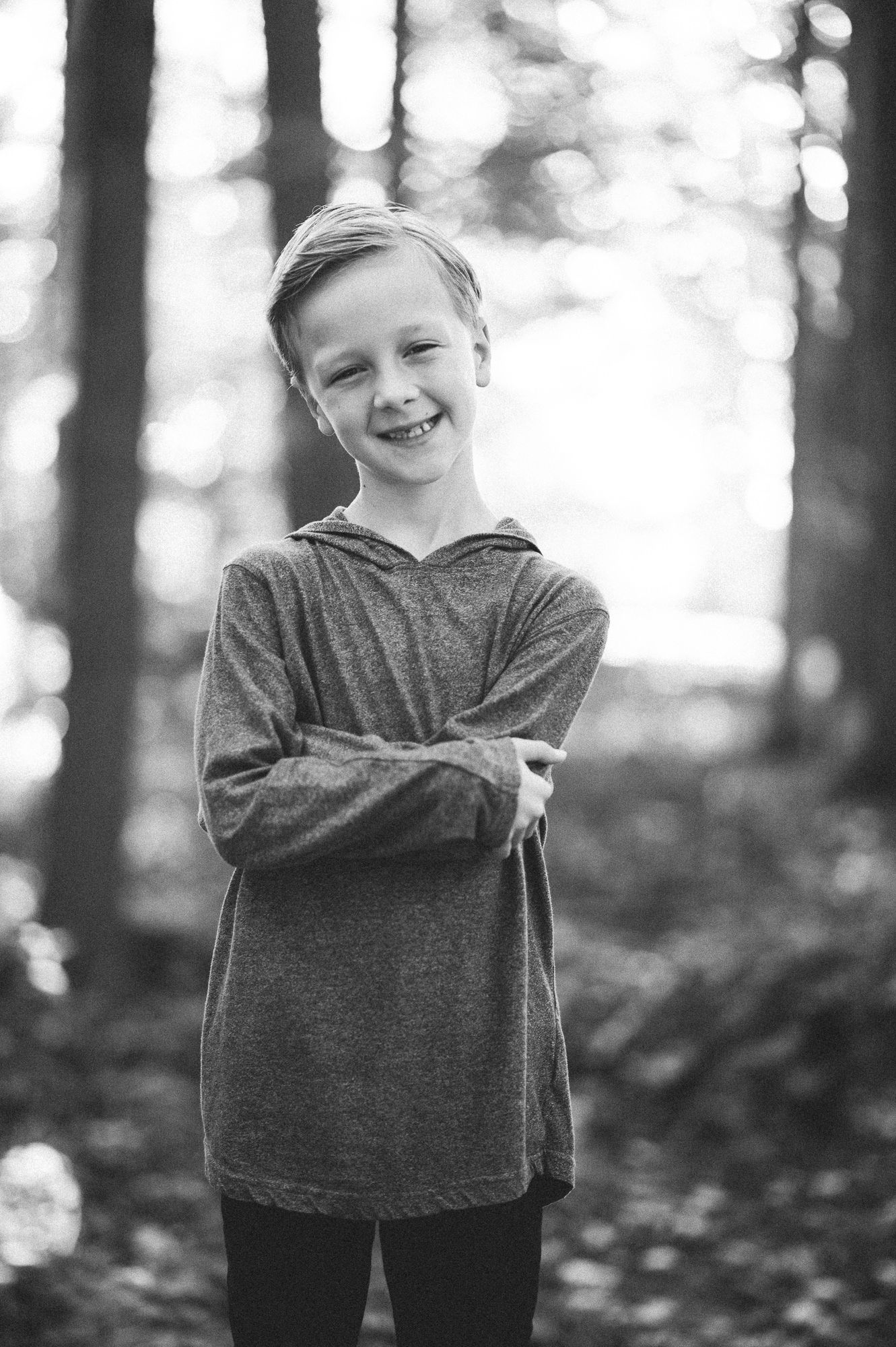 Child in gray sweater poses playfully in a forest setting with bokeh lighting effects.
