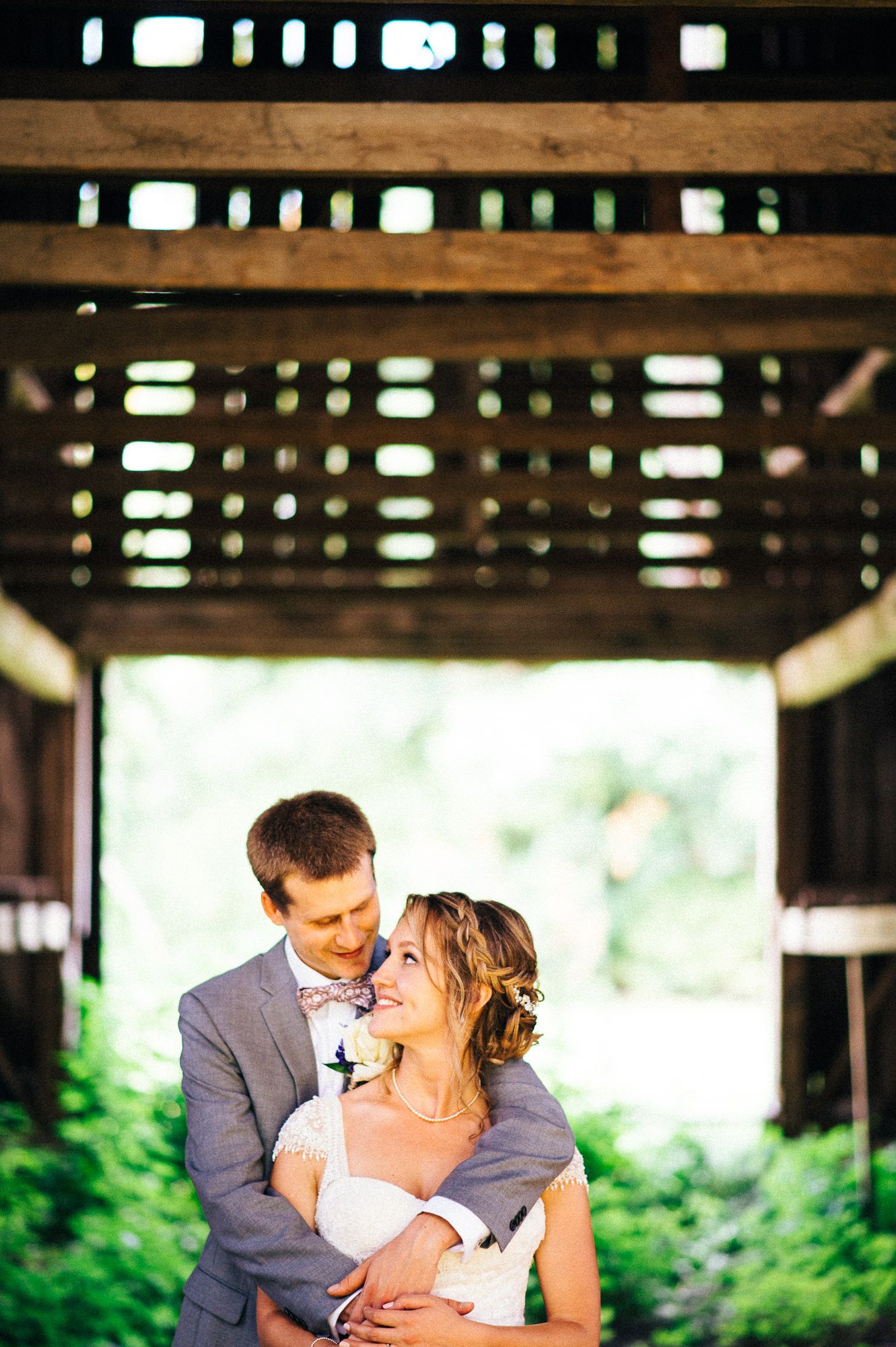 Romantic wedding couple embracing under a rustic wooden pergola surrounded by greenery.