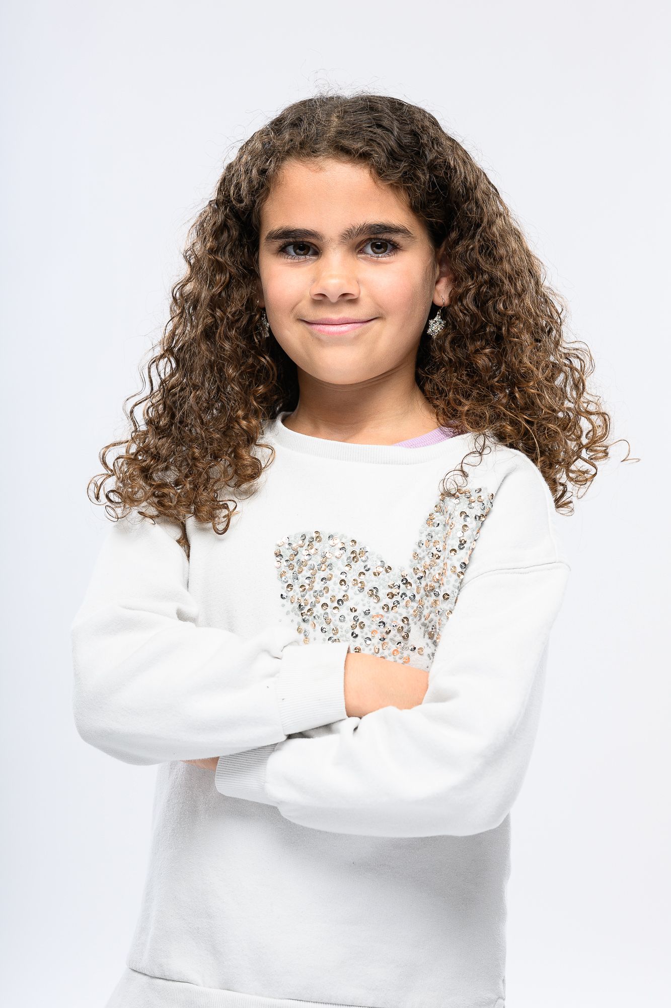 Child with curly hair wearing white sweater poses confidently against white background.