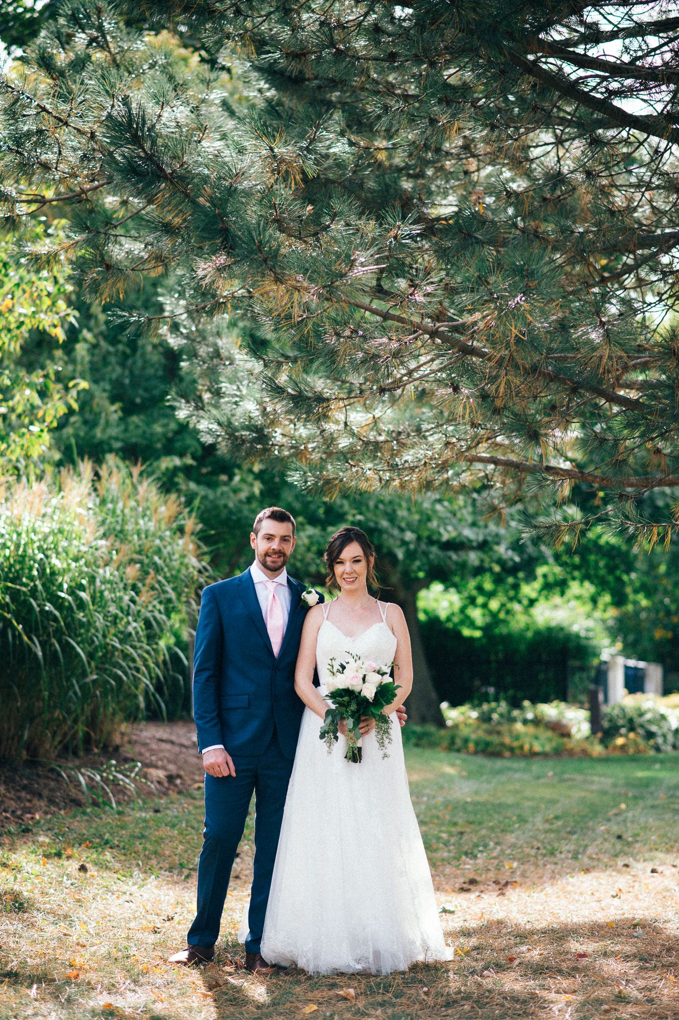 A couple poses under evergreen trees in formal wedding attire with bouquet.