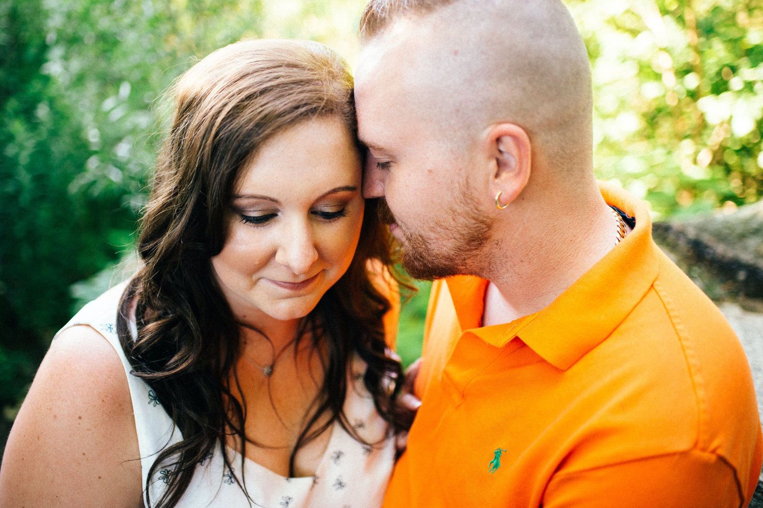 Couple embraces in a sun-dappled forest setting wearing casual attire.