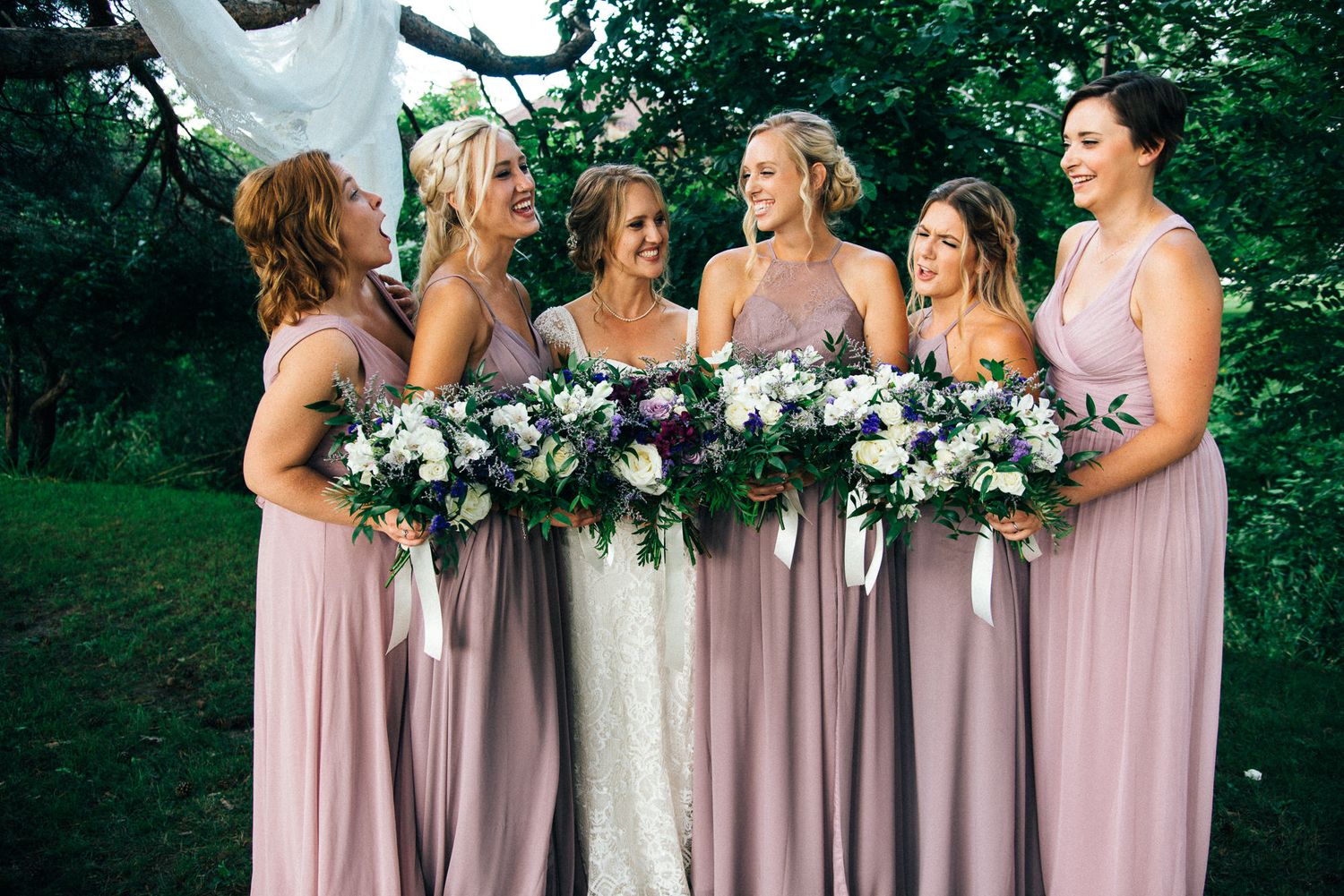 Bridesmaids in blush pink dresses holding floral bouquets pose together outdoors.