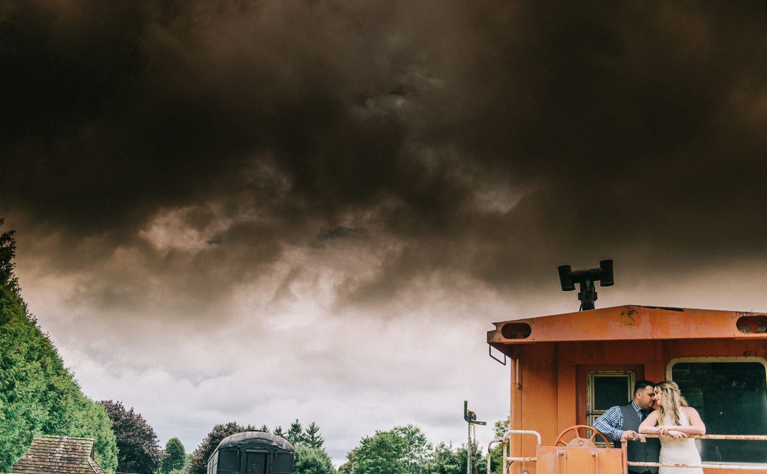 Wedding couple posing on vintage train car under stormy skies.