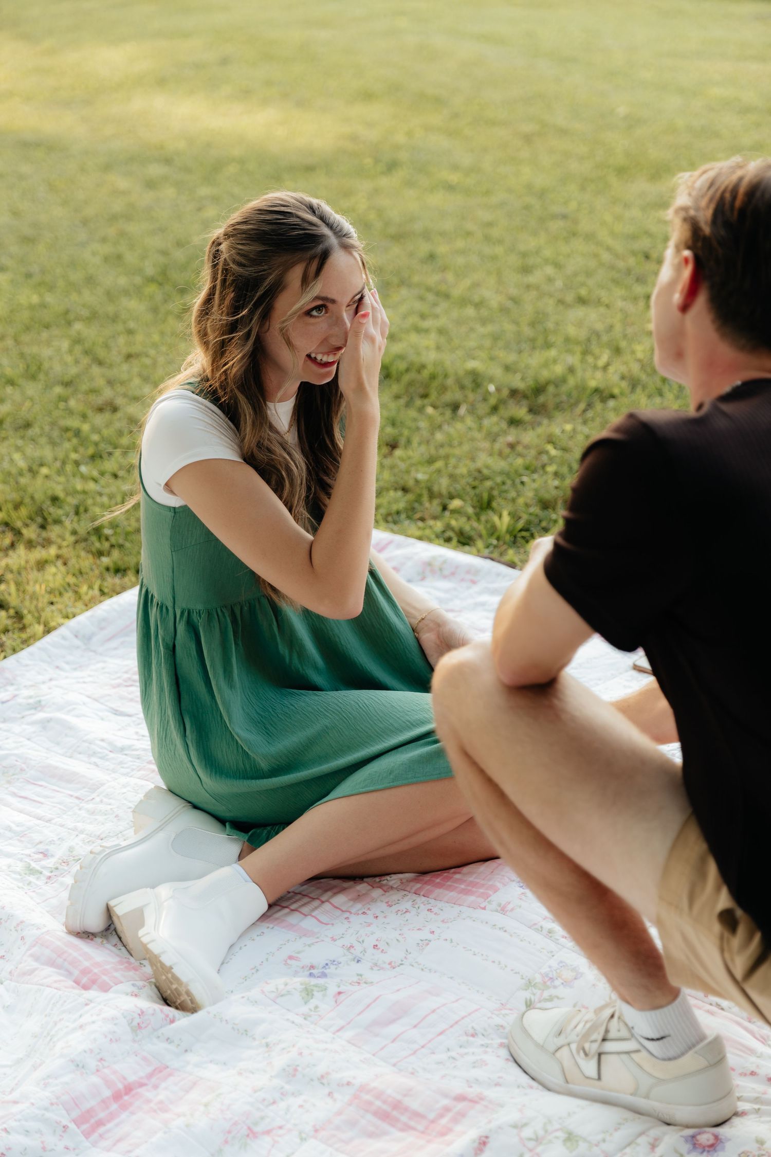 Woman in green dress smiling and sitting on a floral quilt in a grassy park with her partner.