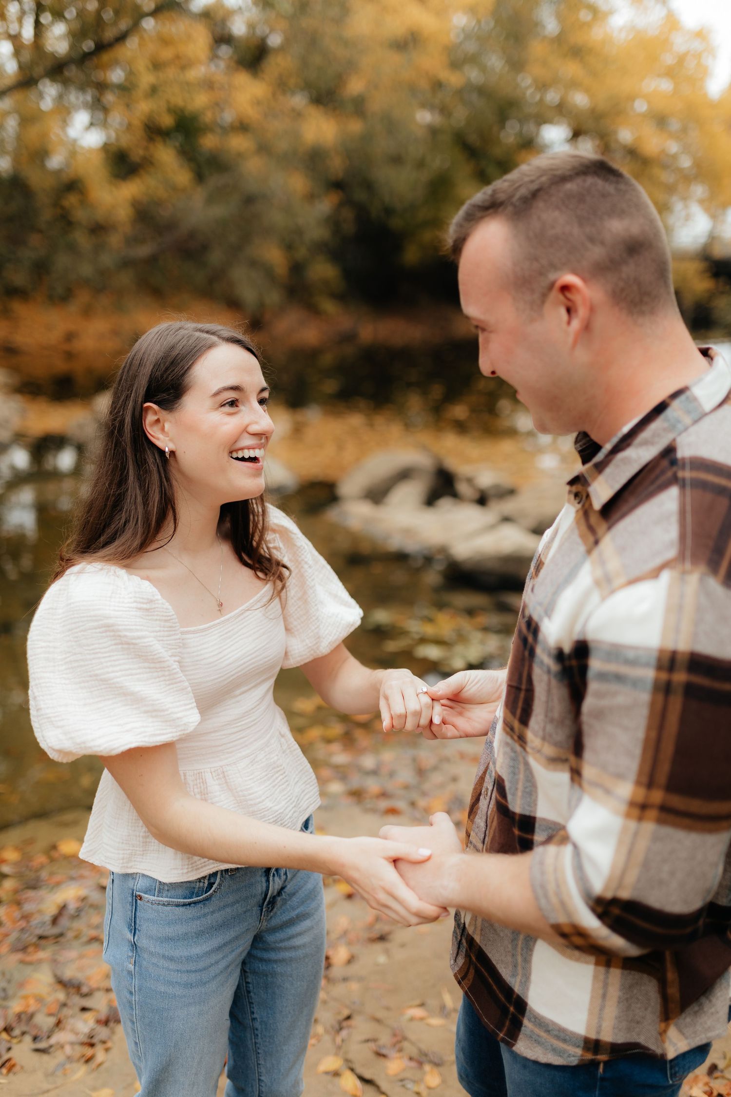 Joyful couple sharing a romantic moment by a rocky autumn creek, woman laughing as man places ring on finger.