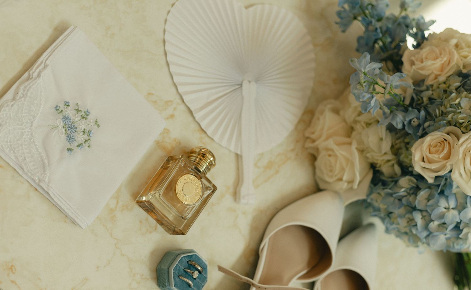 Elegant wedding flat lay with white fan, perfume, blue hydrangea bouquet, heels, and embroidered handkerchief.