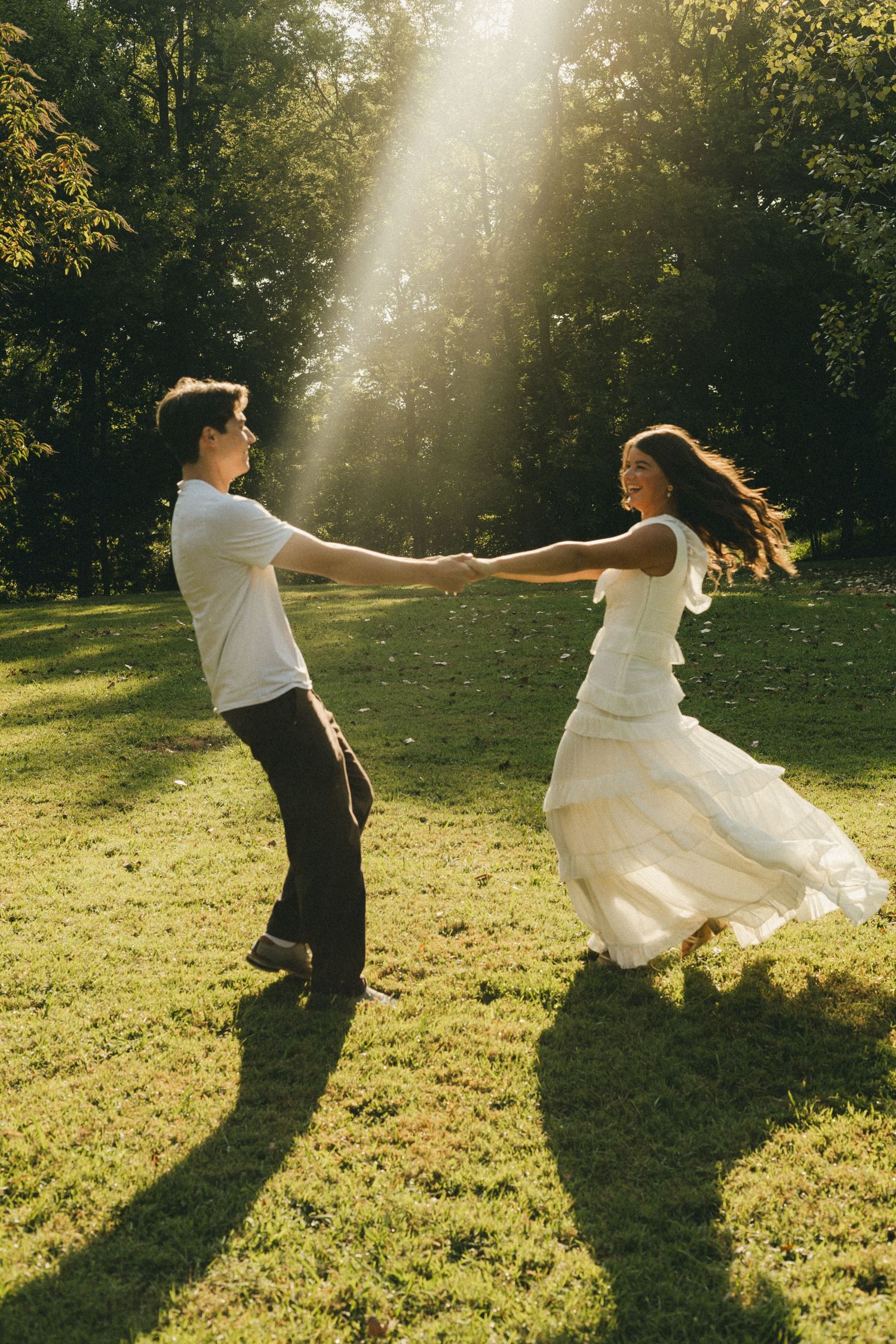 Couple dancing joyfully in sunlit park, woman in flowing white dress, golden hour light streaming through green trees.