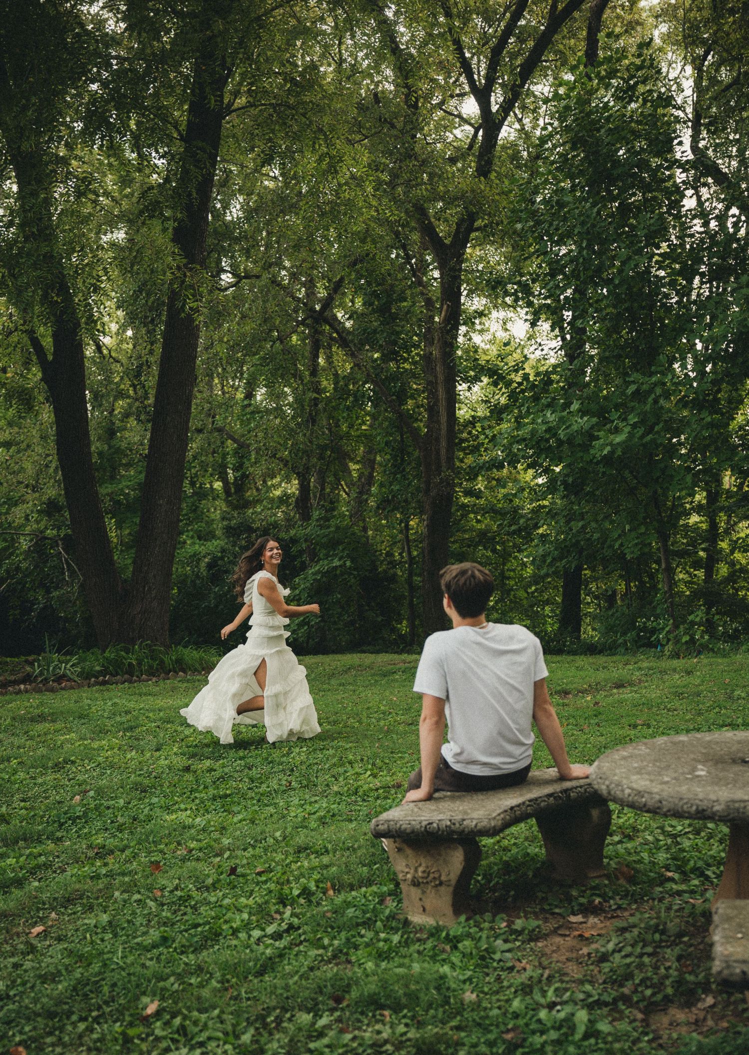 A bride in a white gown runs toward a groom seated on a stone bench in a lush green park.