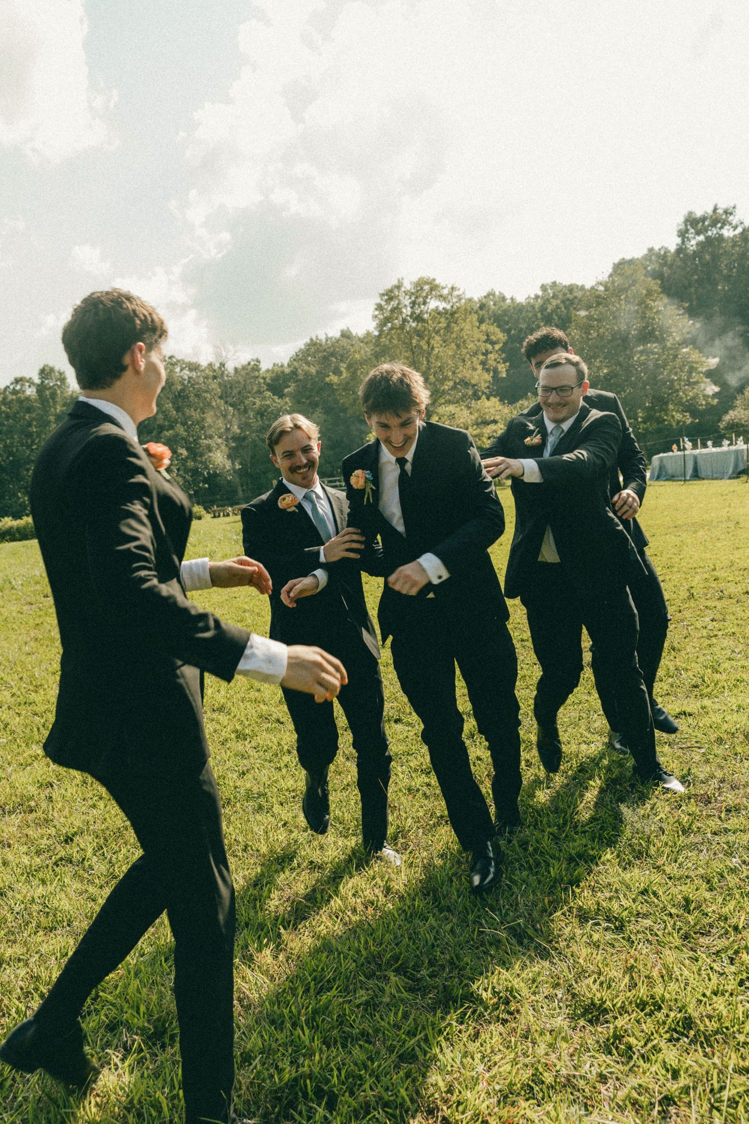 Groomsmen in black suits dancing and celebrating outdoors on green grass at a wedding.