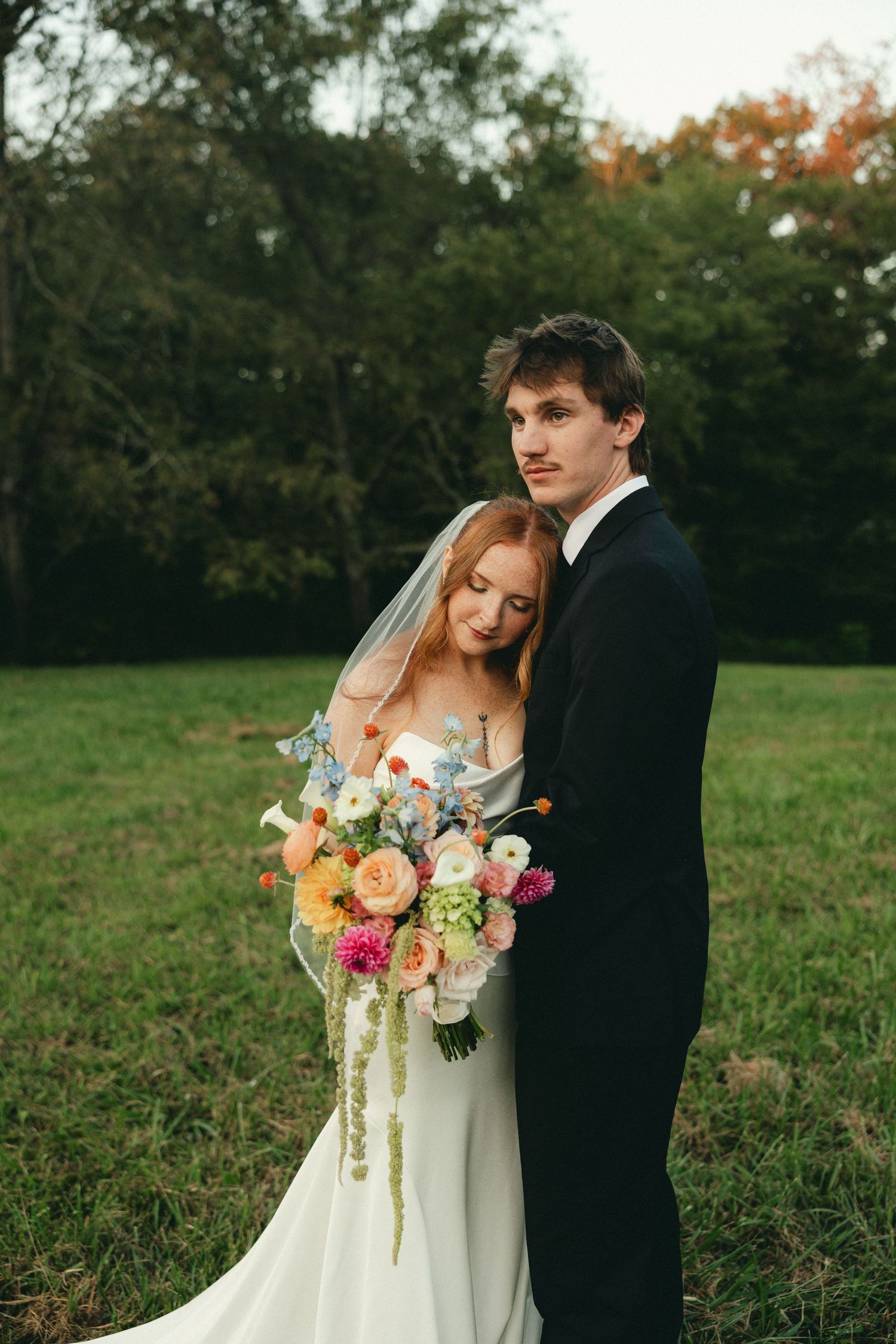 Bride and groom embracing outdoors, bride holding colorful bouquet in white gown with veil, groom in black tuxedo.