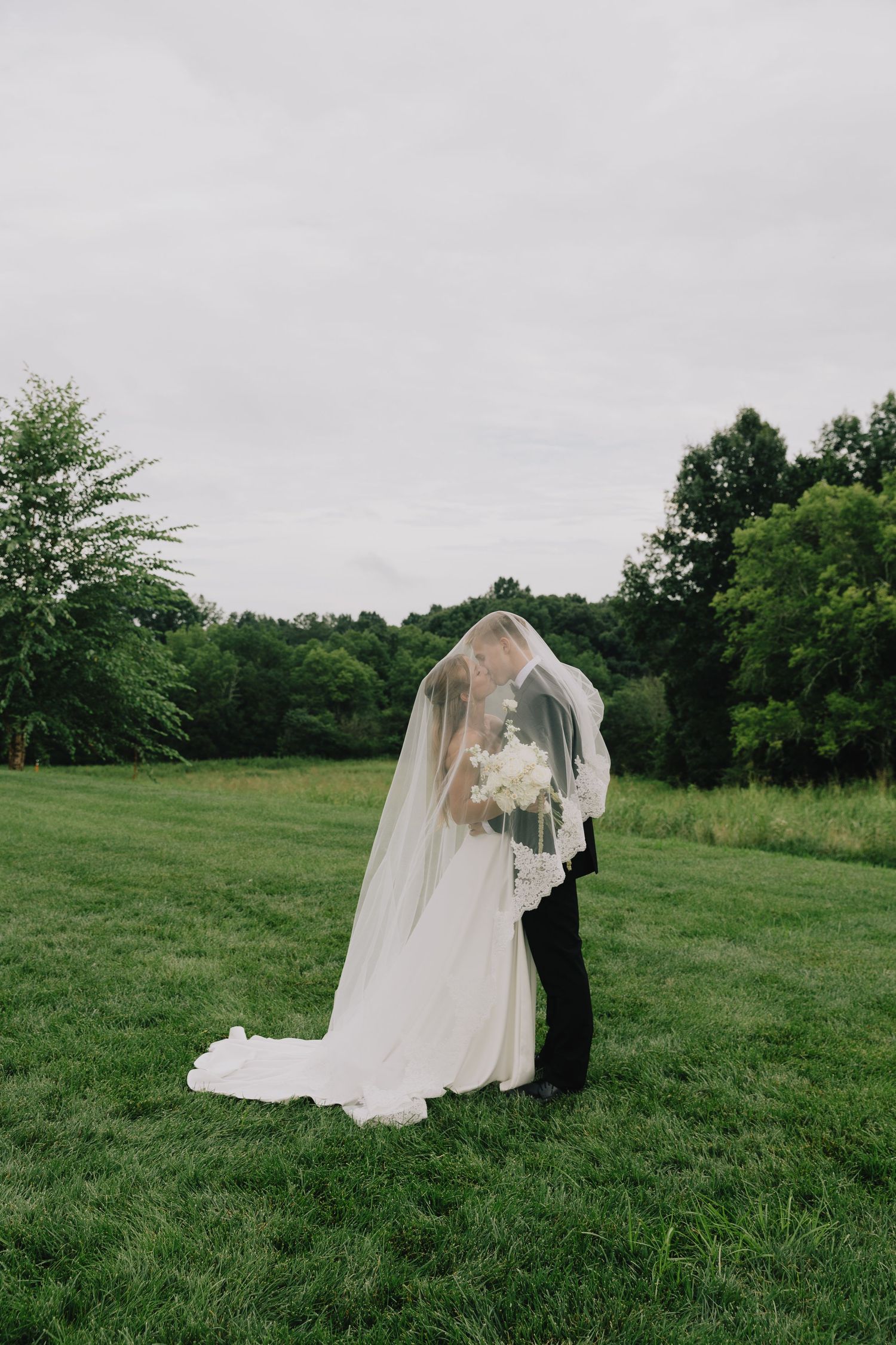 Bride and groom share a kiss under a flowing white veil on a lush green lawn.