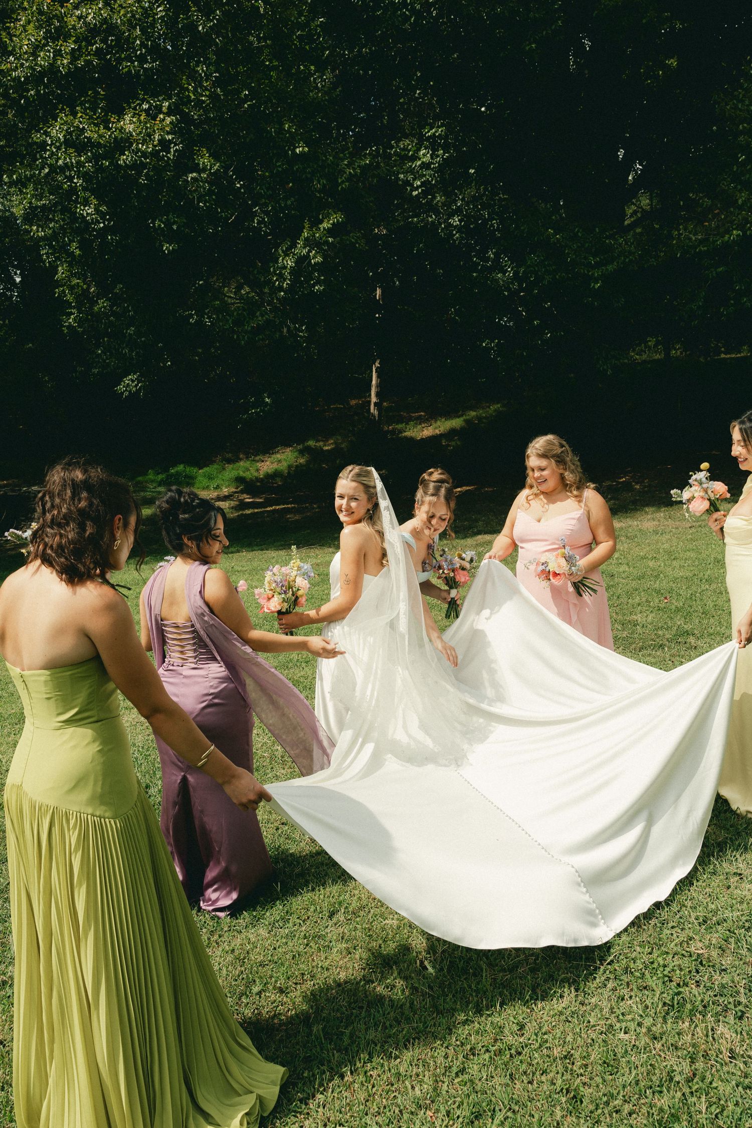 Bride in white gown with bridesmaids in colorful dresses spreading her train on green lawn outdoors.