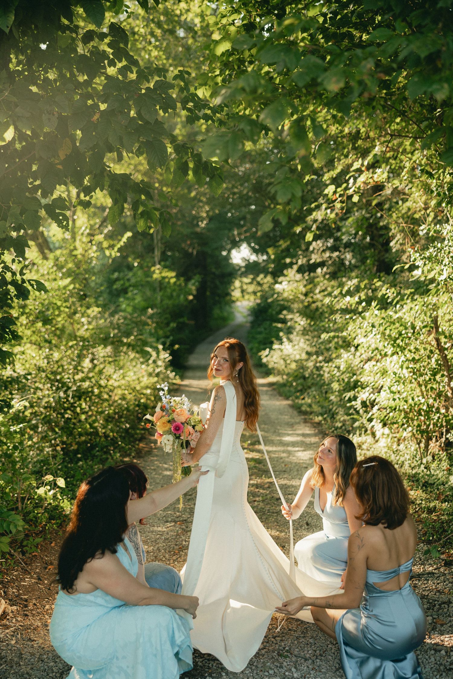 Bride in white gown holds bouquet while bridesmaids in blue dresses arrange her train on a sunlit tree-lined path.