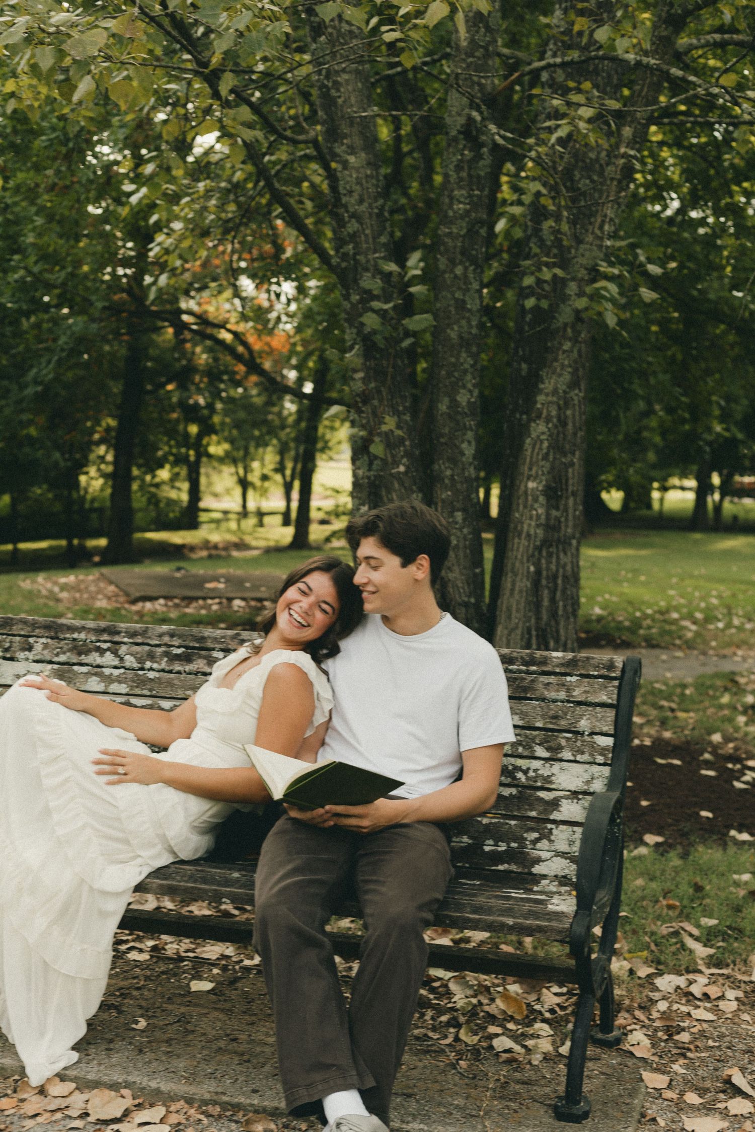 A smiling couple sits on a park bench, woman in white dress leaning on man holding a book, surrounded by green trees.