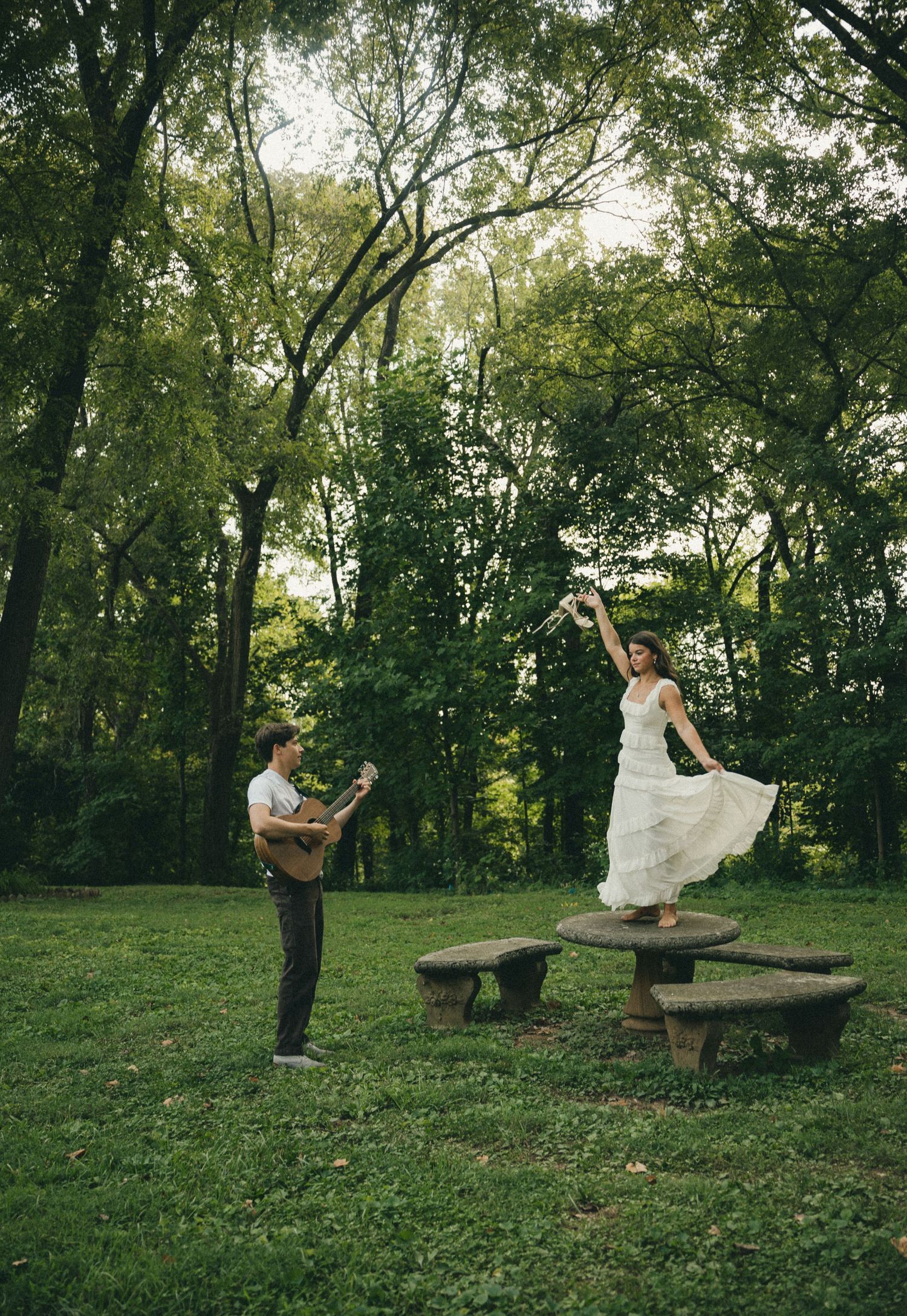 Bride in white gown dancing on picnic table while groom plays guitar in lush green park.