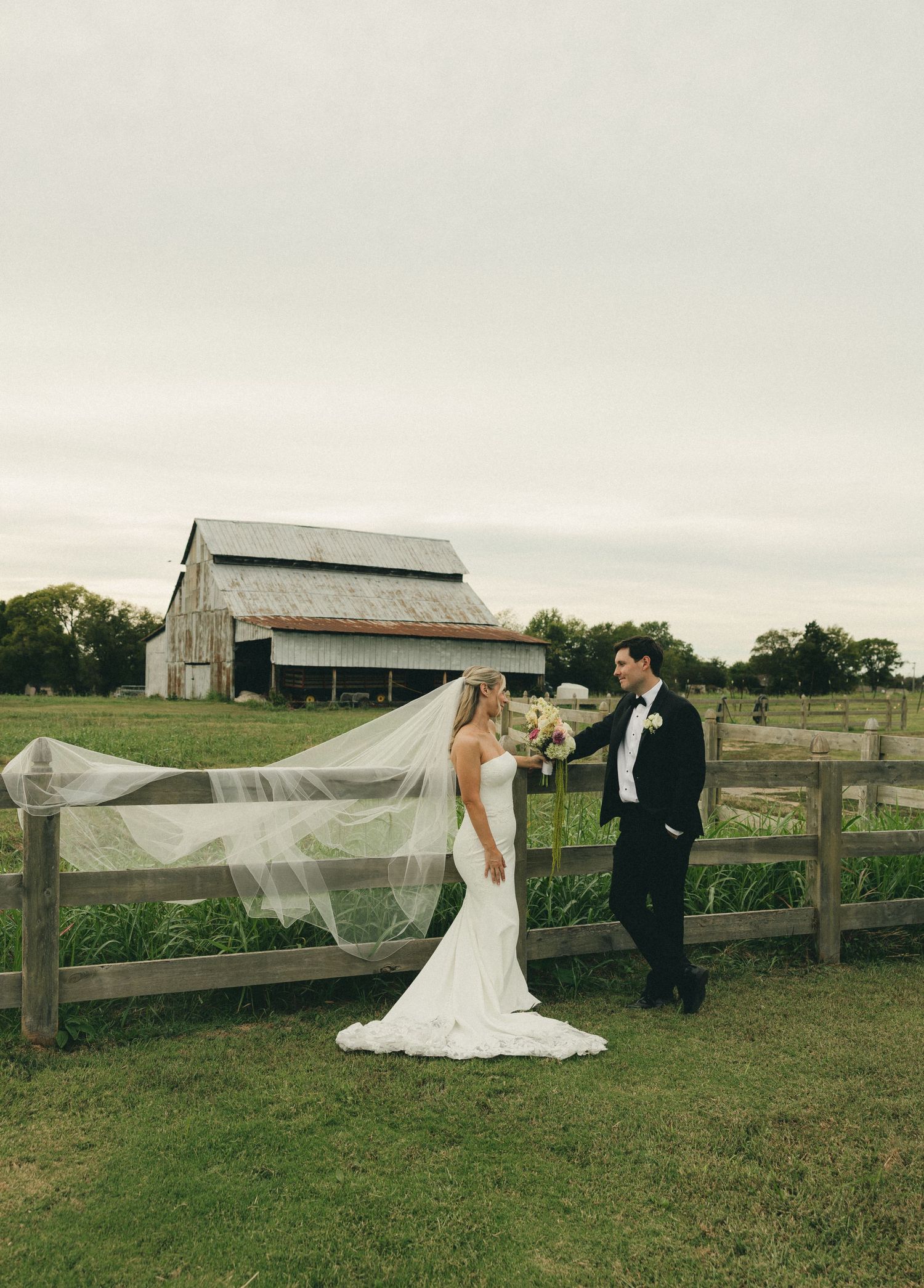 Bride and groom pose by rustic wooden fence with barn backdrop, cathedral veil flowing in the wind.