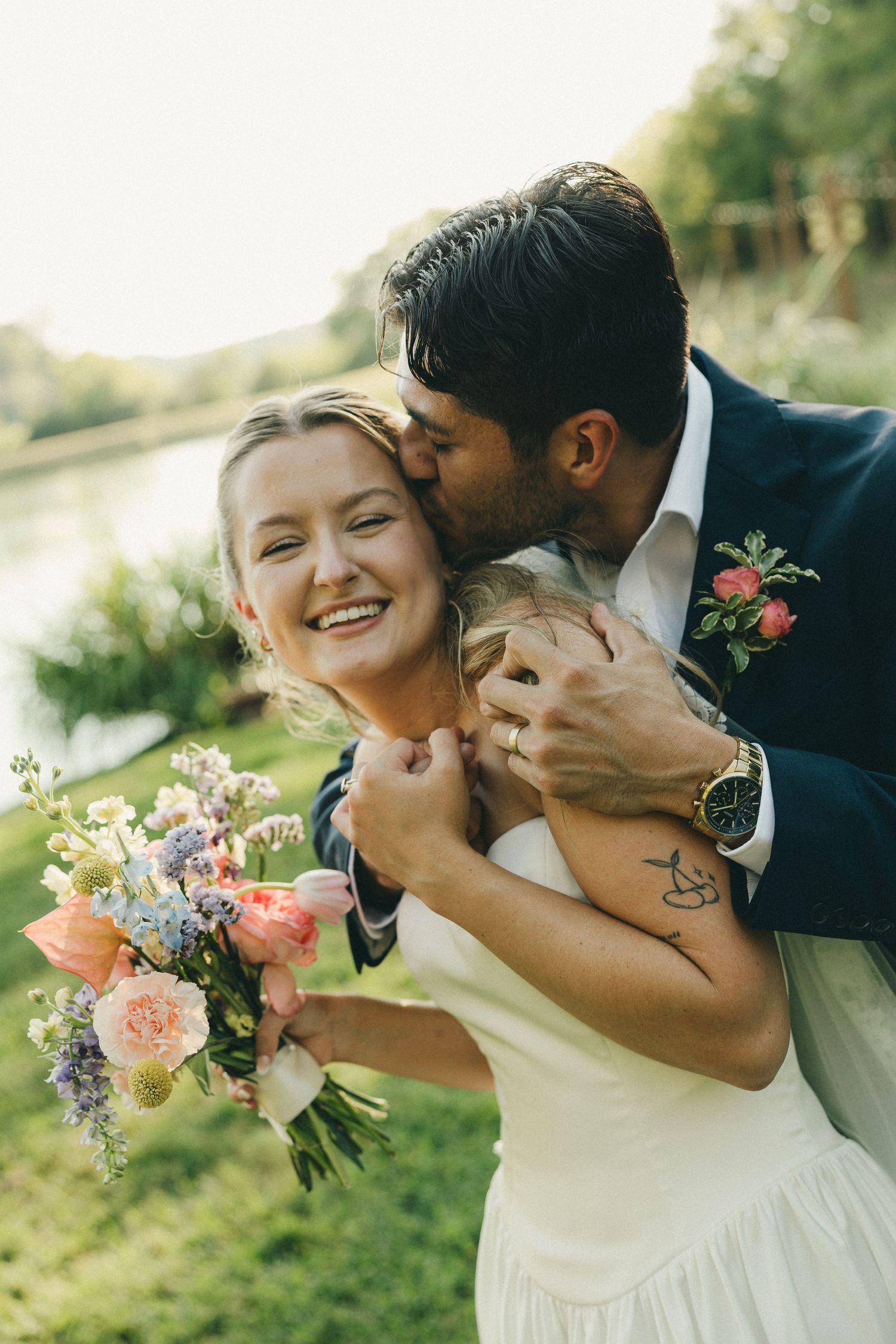 Groom in navy suit kisses smiling bride holding colorful floral bouquet outdoors near a lake on their wedding day.
