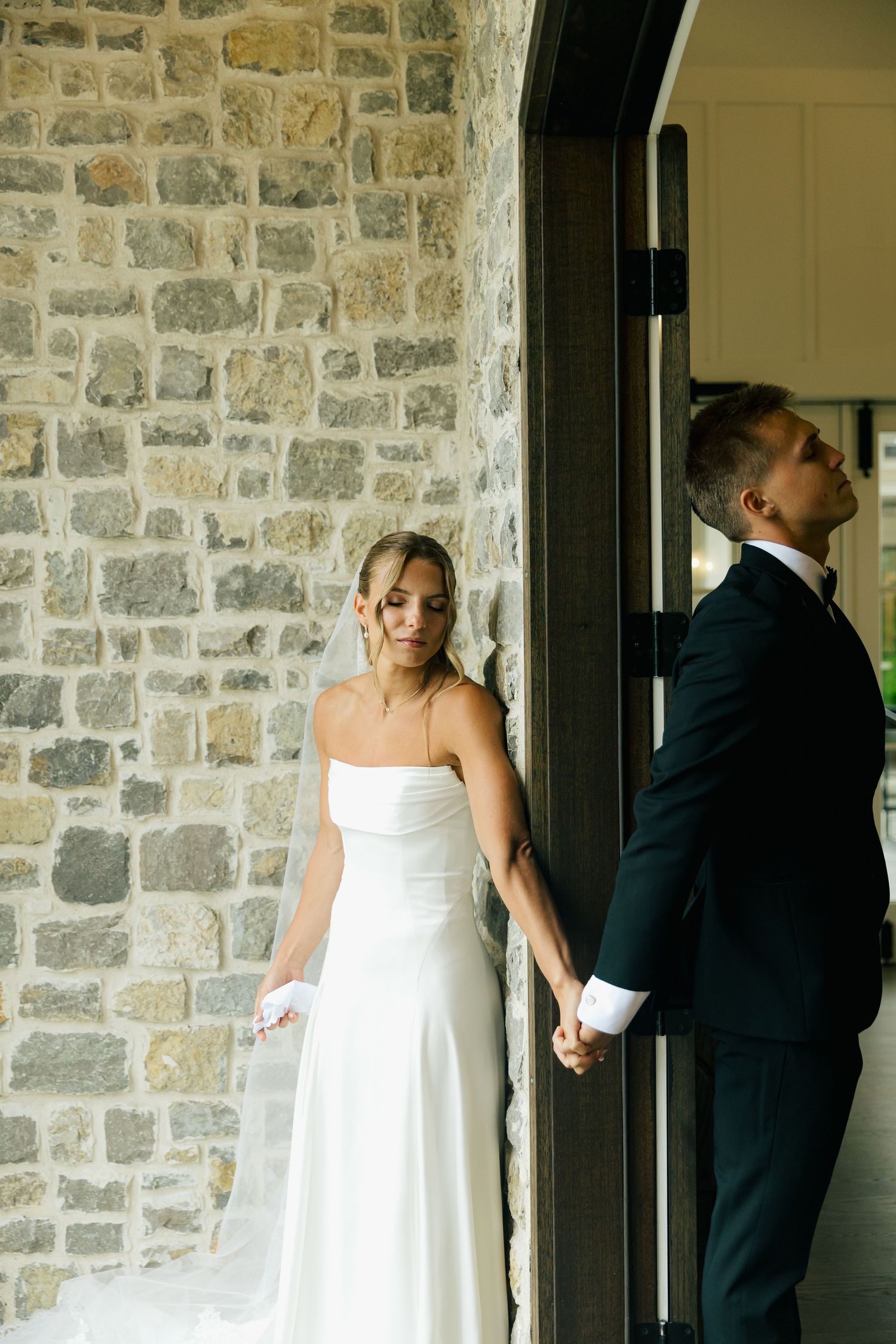 Bride in white strapless gown and groom in black suit hold hands around a stone pillar at their wedding.