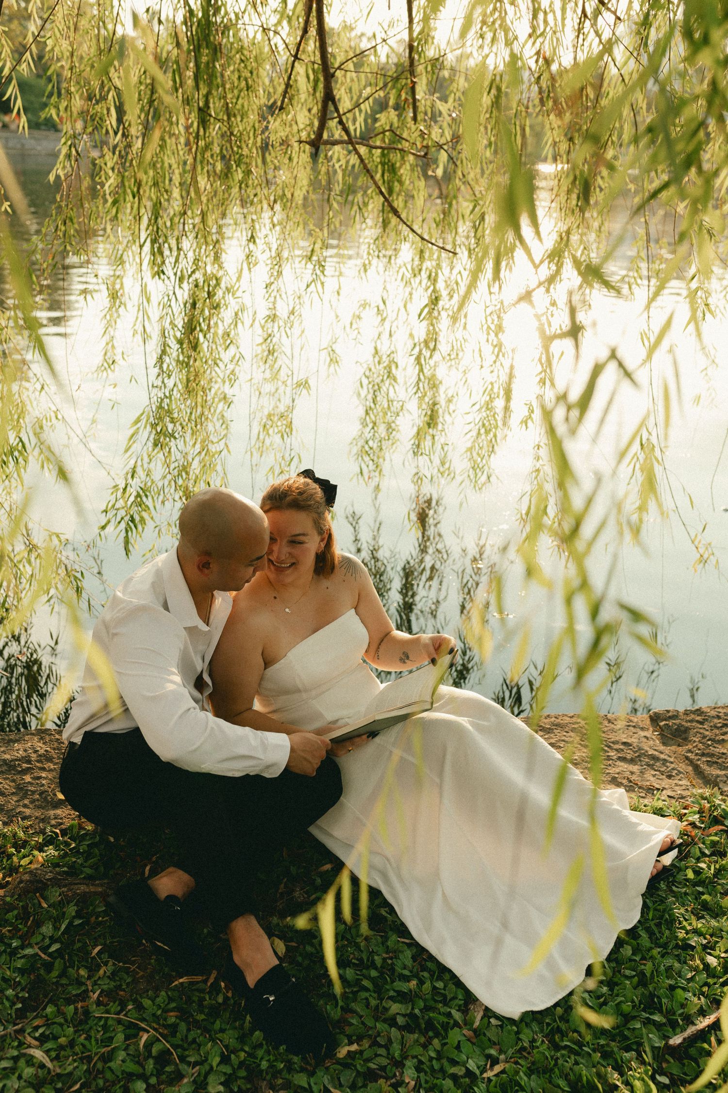 Bride and groom sit under weeping willow by lake, sharing a book in wedding attire.