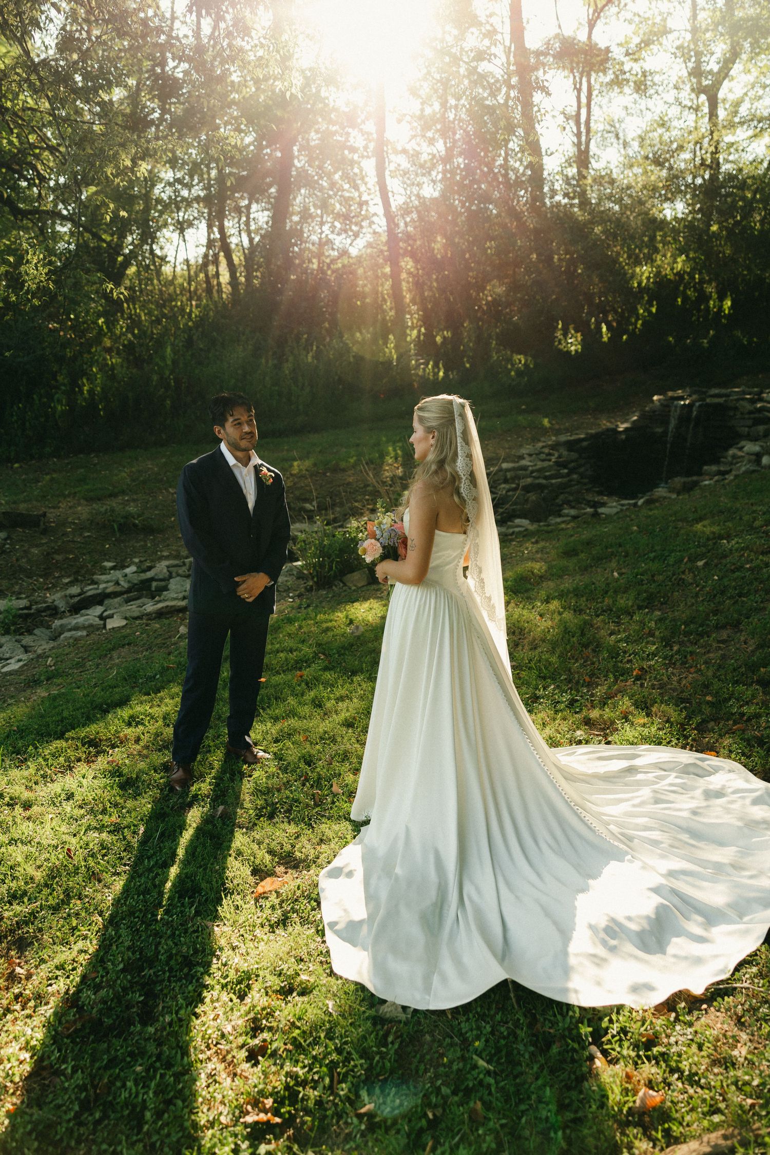Bride in white gown with veil holds bouquet facing groom in black suit outdoors in sunlit garden setting.