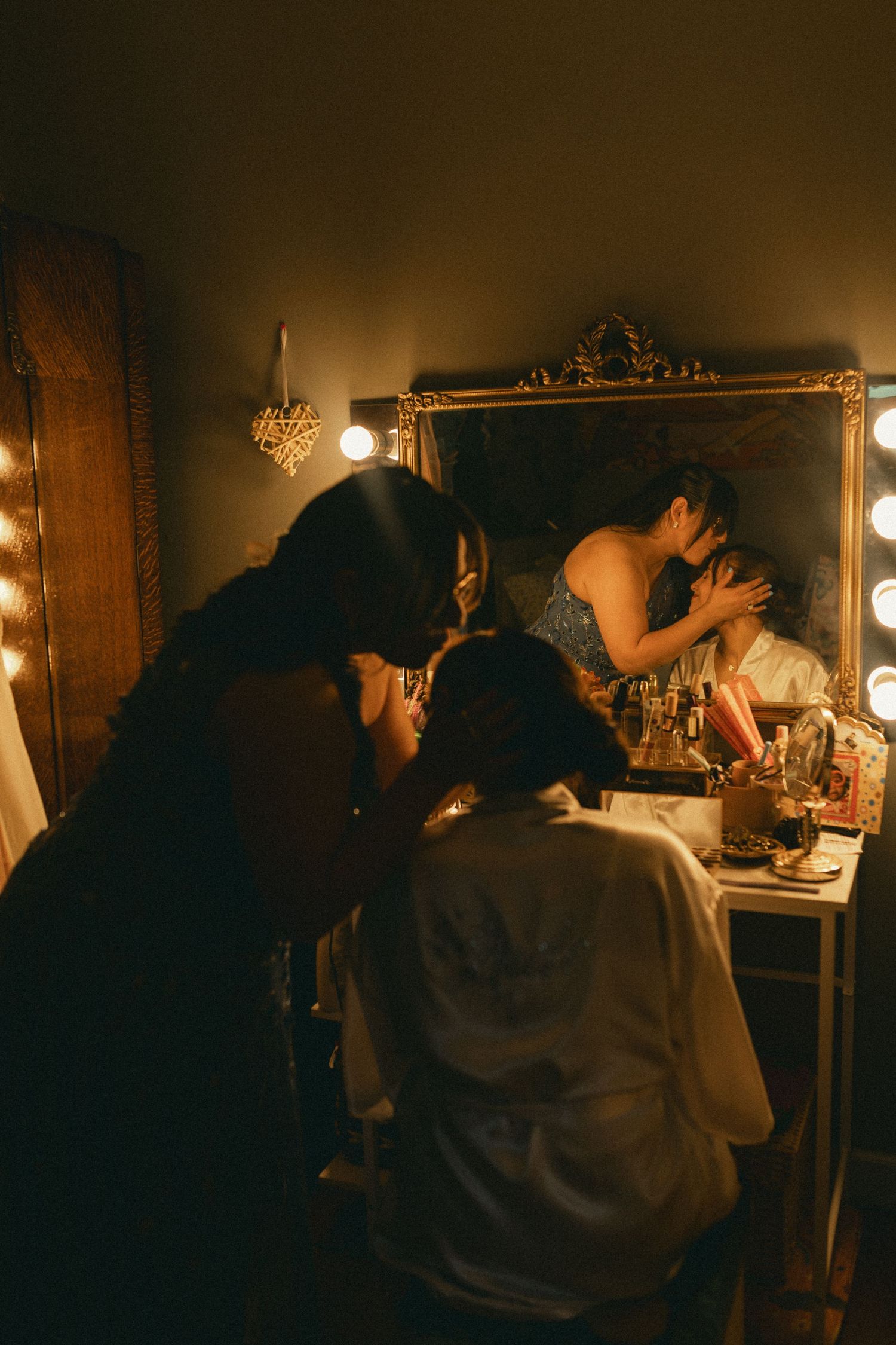 Bride getting makeup done at lit vanity mirror in dimly lit bridal preparation room.