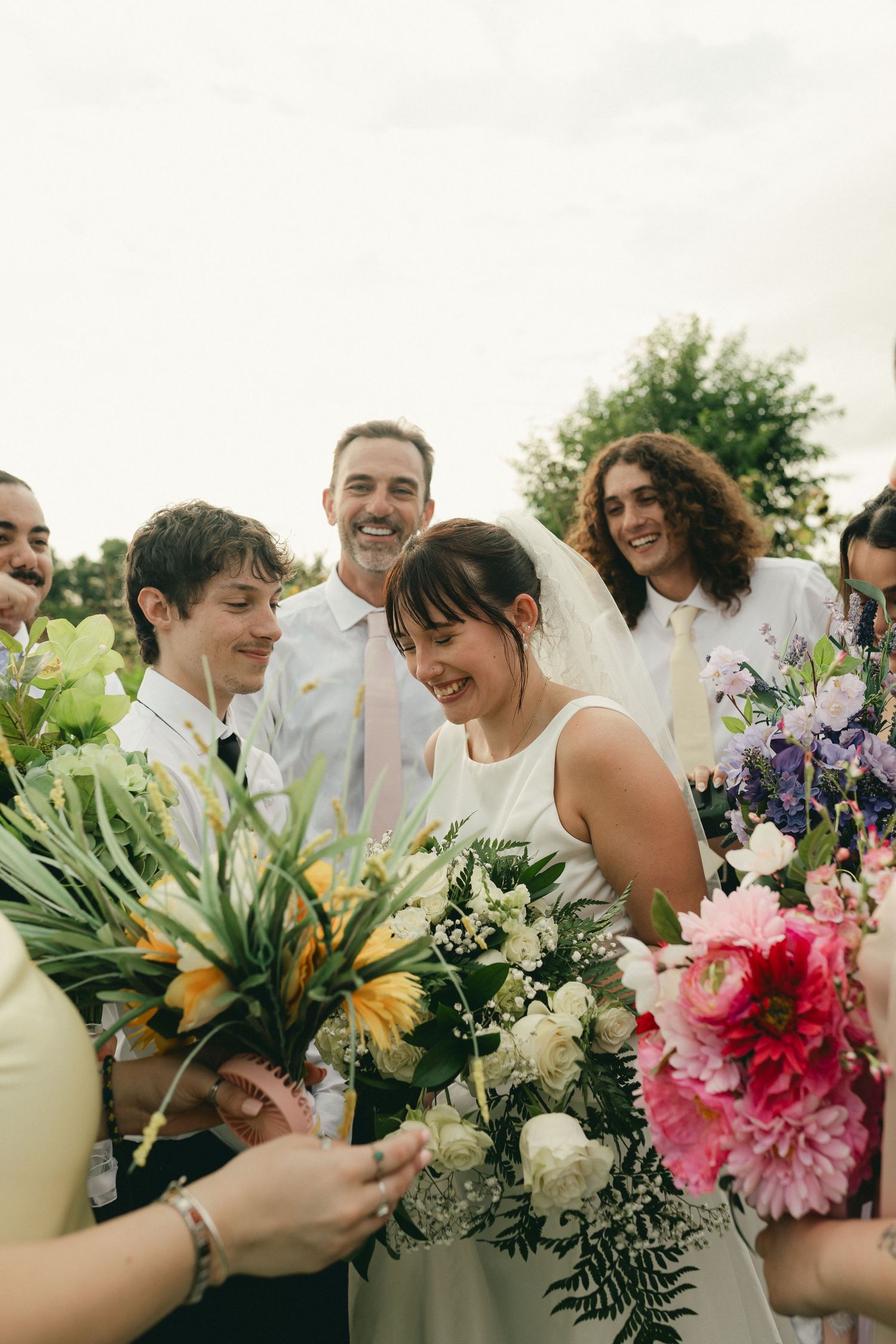 Bride laughing with wedding guests while holding floral bouquets in outdoor setting.