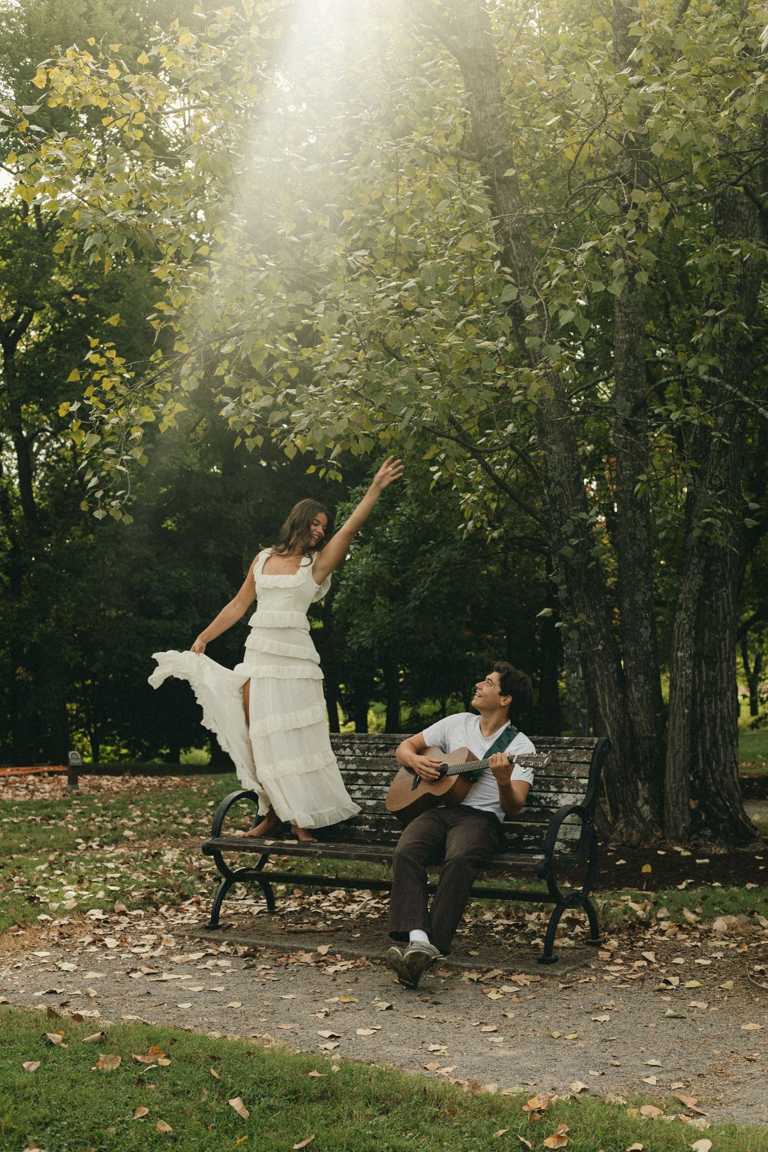 Couple in a sunlit park, woman in white dress dancing on bench while man plays acoustic guitar.