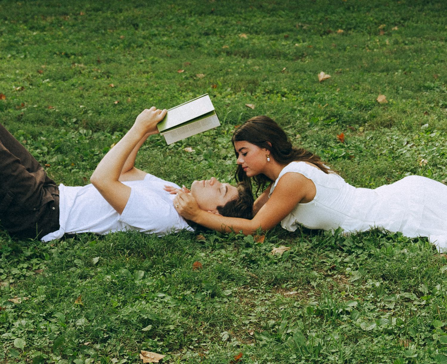 Couple lying on green grass, woman in white dress leaning toward man reading a book outdoors.