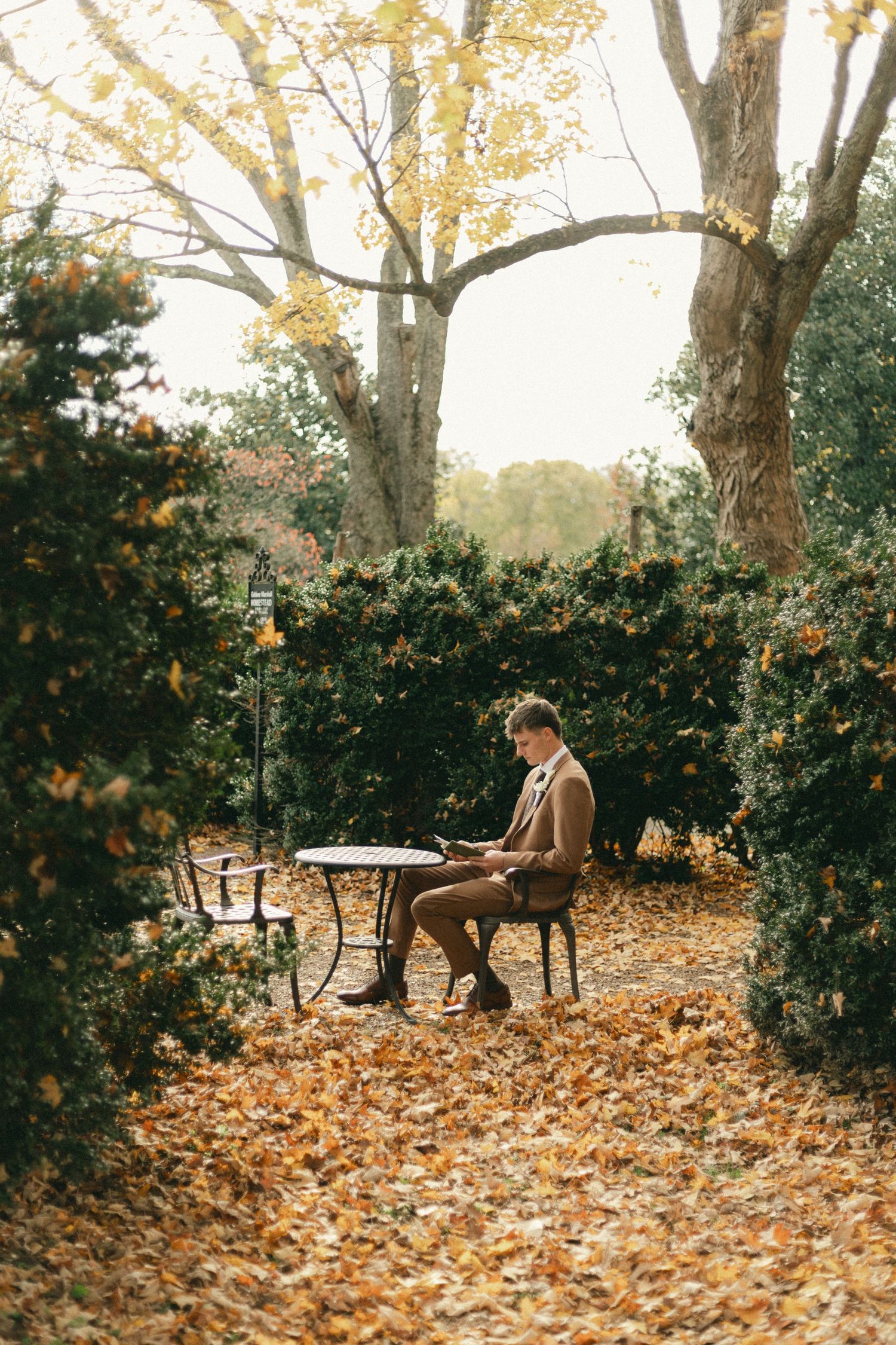 Man in tan suit reading at ornate table surrounded by autumn leaves and green hedges in a serene fall garden.