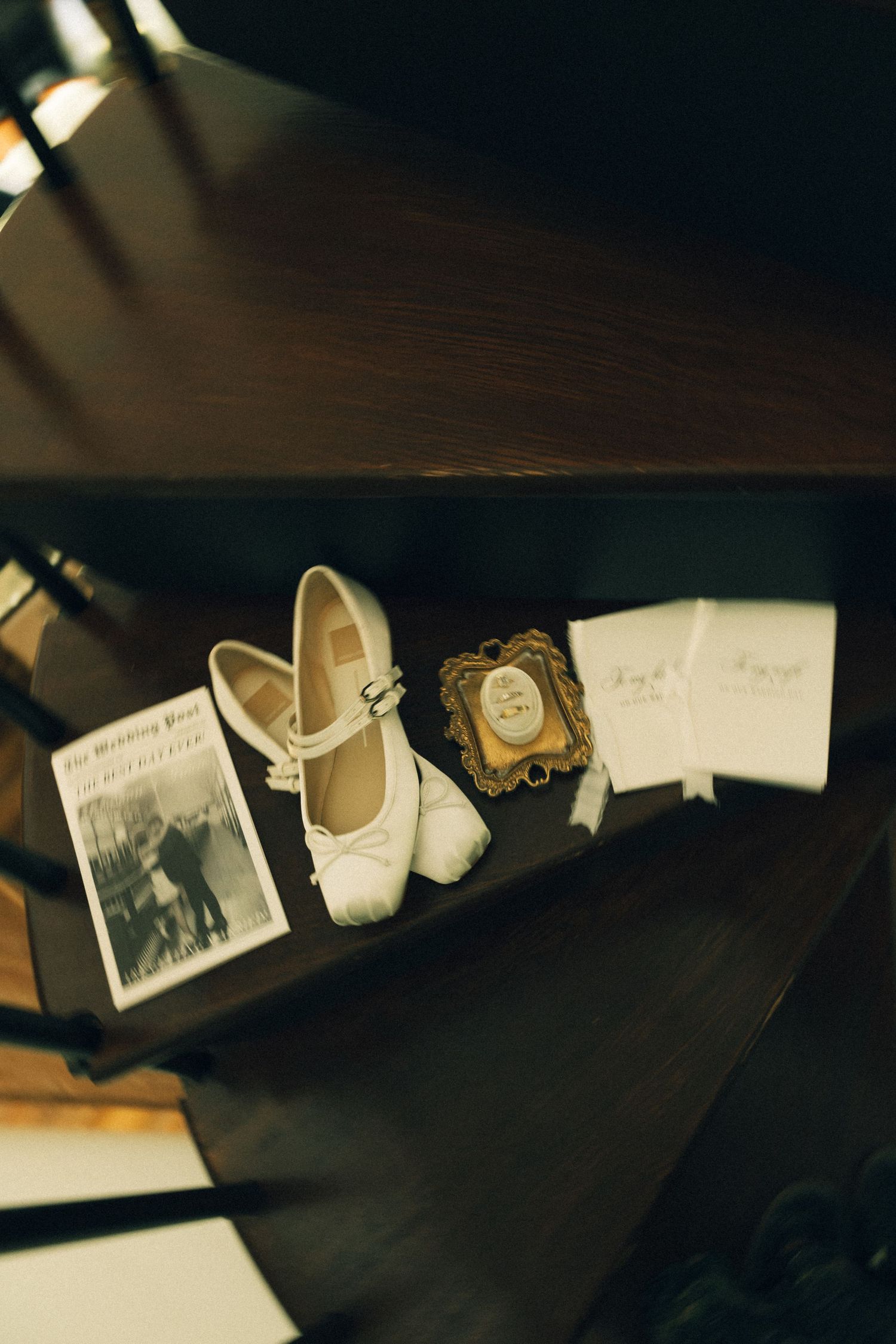 White bridal heels displayed on dark wood shelf alongside vintage book, ornate frame, and wedding stationery.