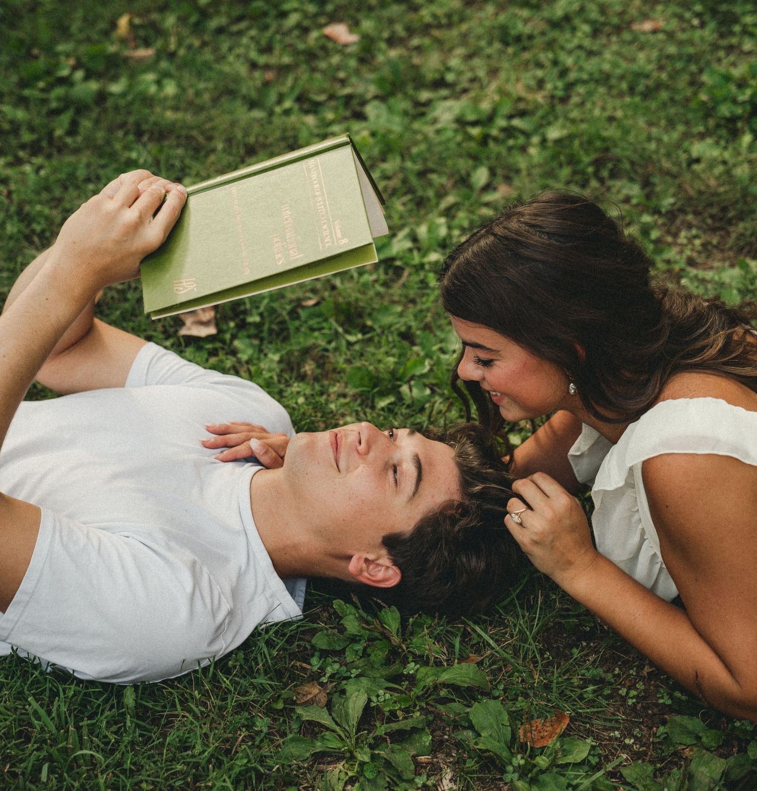 Couple lying on grass in a park, man reading a green book while woman smiles at him romantically.