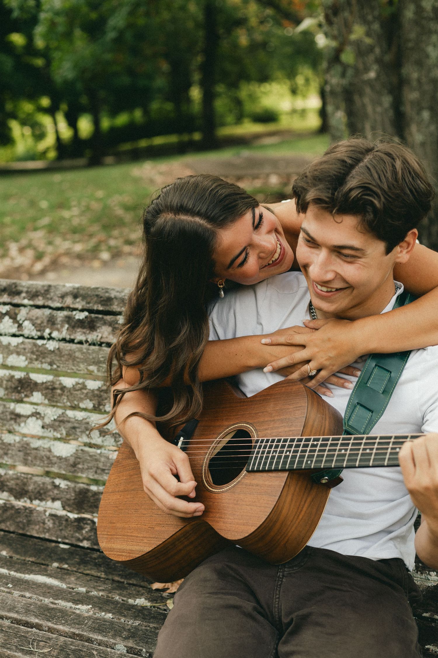 Young couple sitting on a park bench, man playing acoustic guitar while woman hugs him from behind, smiling.