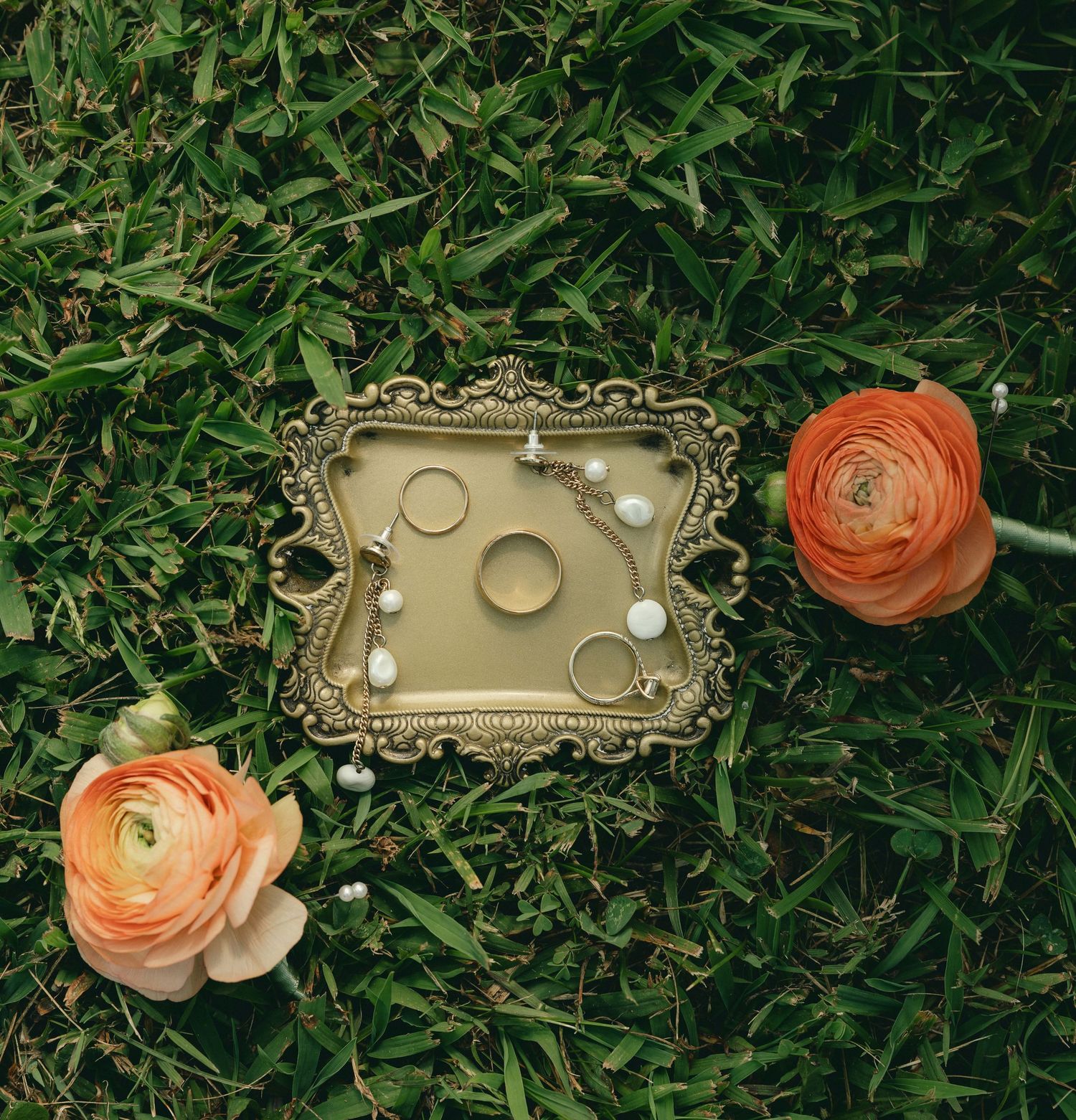 Gold ornate tray with wedding rings and pearl jewelry on green grass beside orange ranunculus flowers.