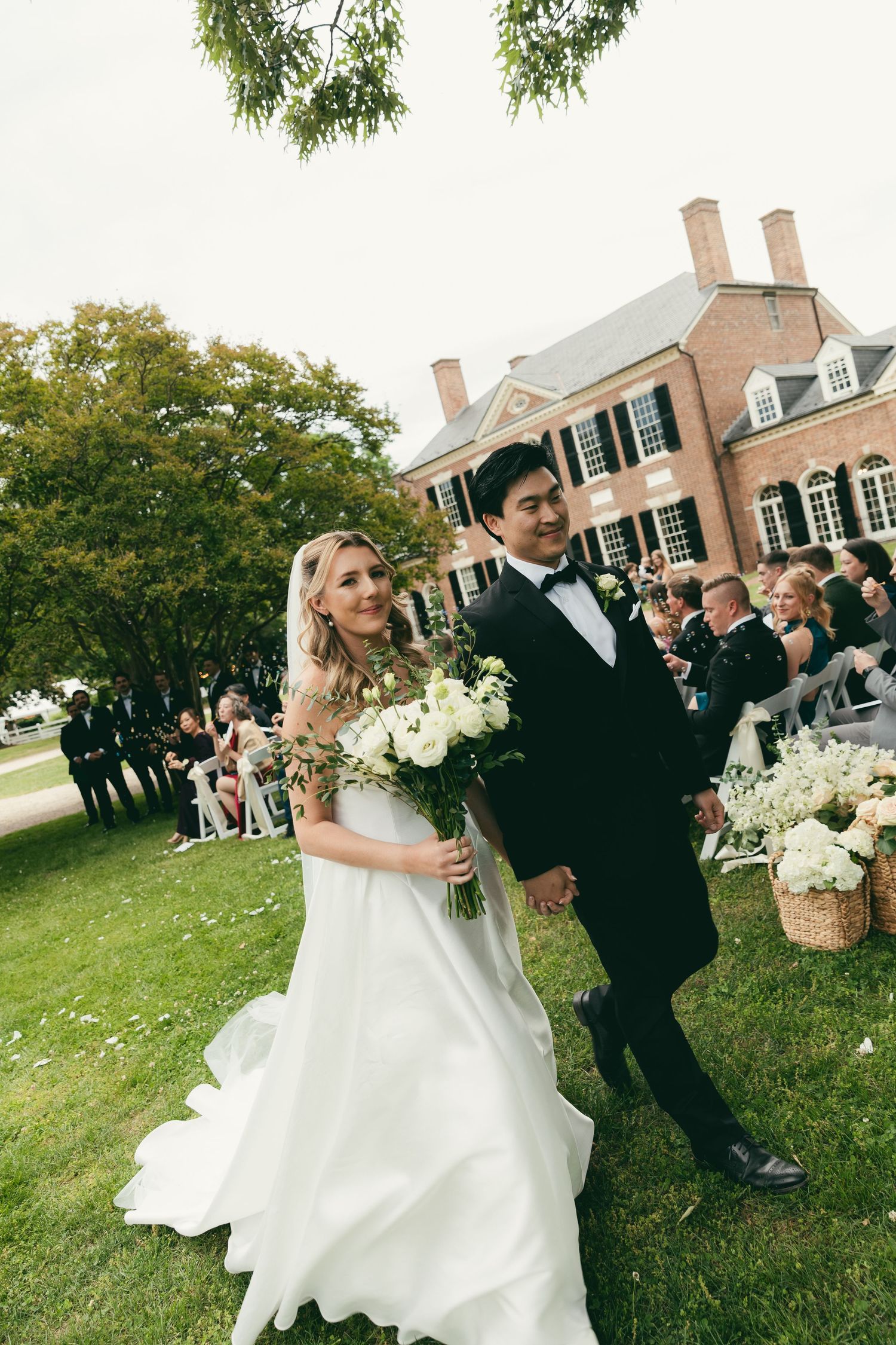 Newlyweds walking hand in hand after outdoor ceremony at elegant brick estate mansion venue.