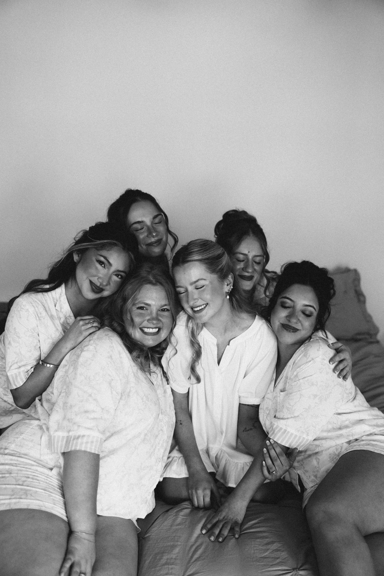 Five women in white robes laughing and posing together on a bed in a black and white photo.
