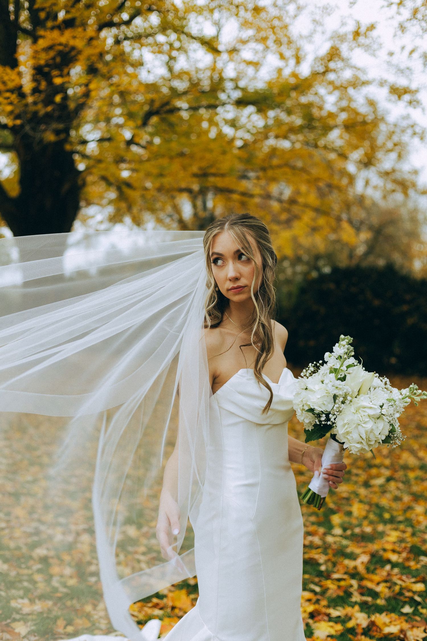 Bride in white gown with flowing veil holding bouquet among autumn yellow leaves.