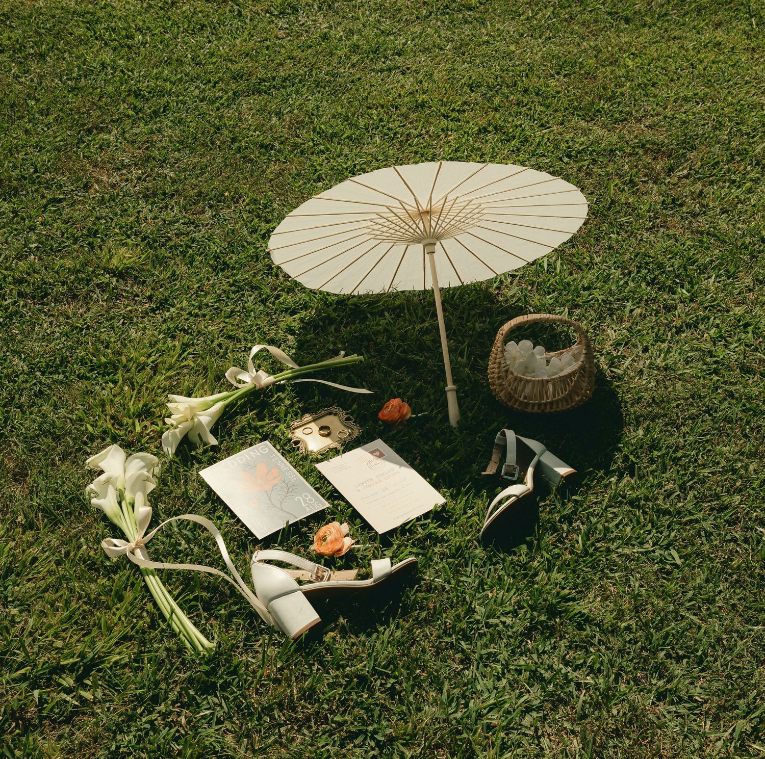 A white paper parasol surrounded by flowers, books, a wicker basket, and elegant accessories on lush green grass.