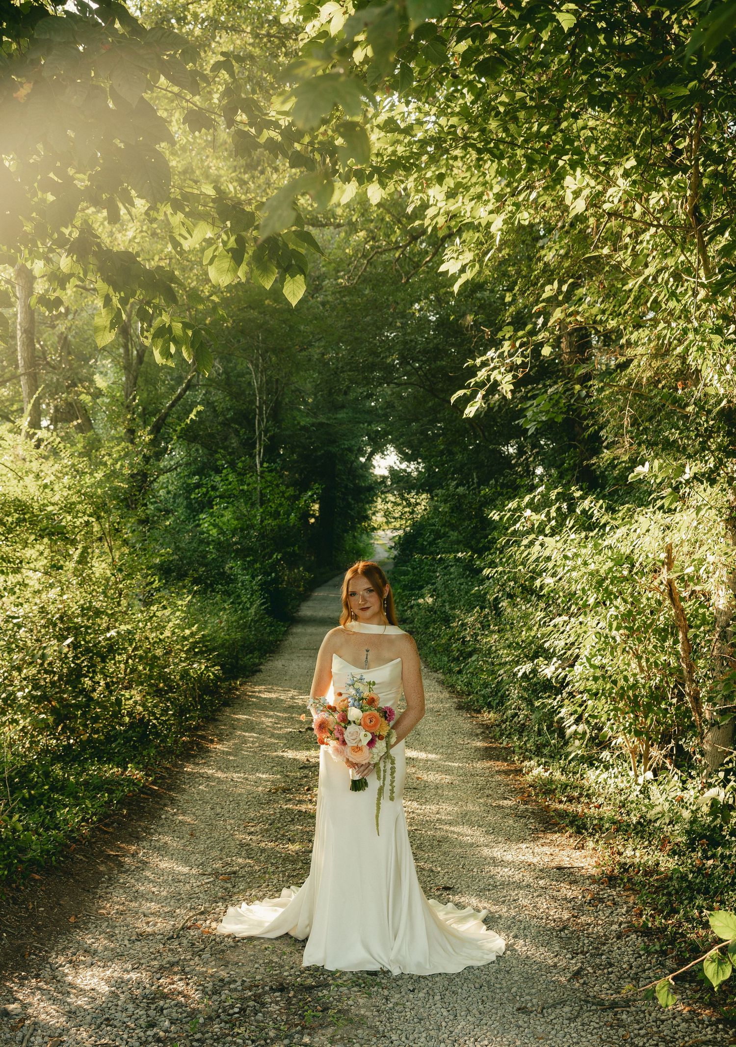 Bride in white gown holding colorful bouquet stands on tree-lined gravel path in golden sunlight.