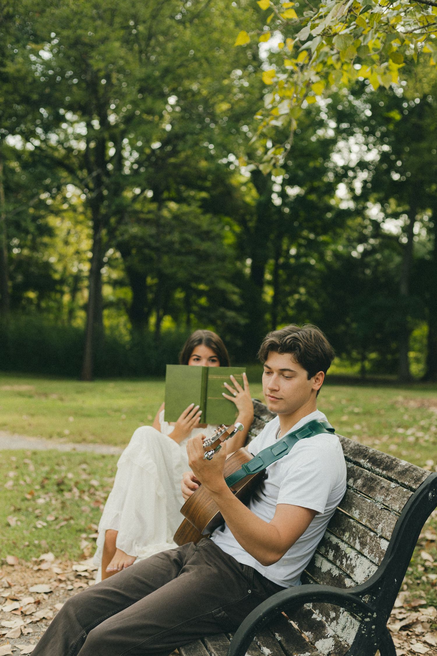 Young man playing acoustic guitar on a park bench while woman reads a green book outdoors.