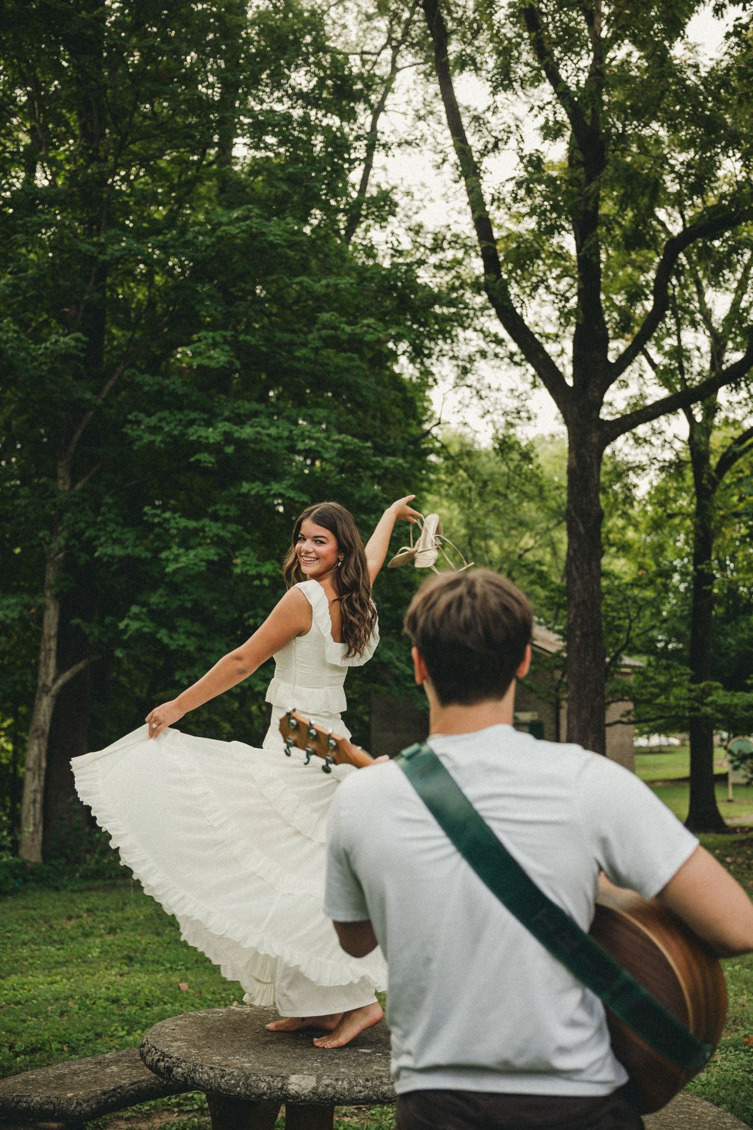 Bride in white dress twirling while groom plays acoustic guitar outdoors in lush green park setting.