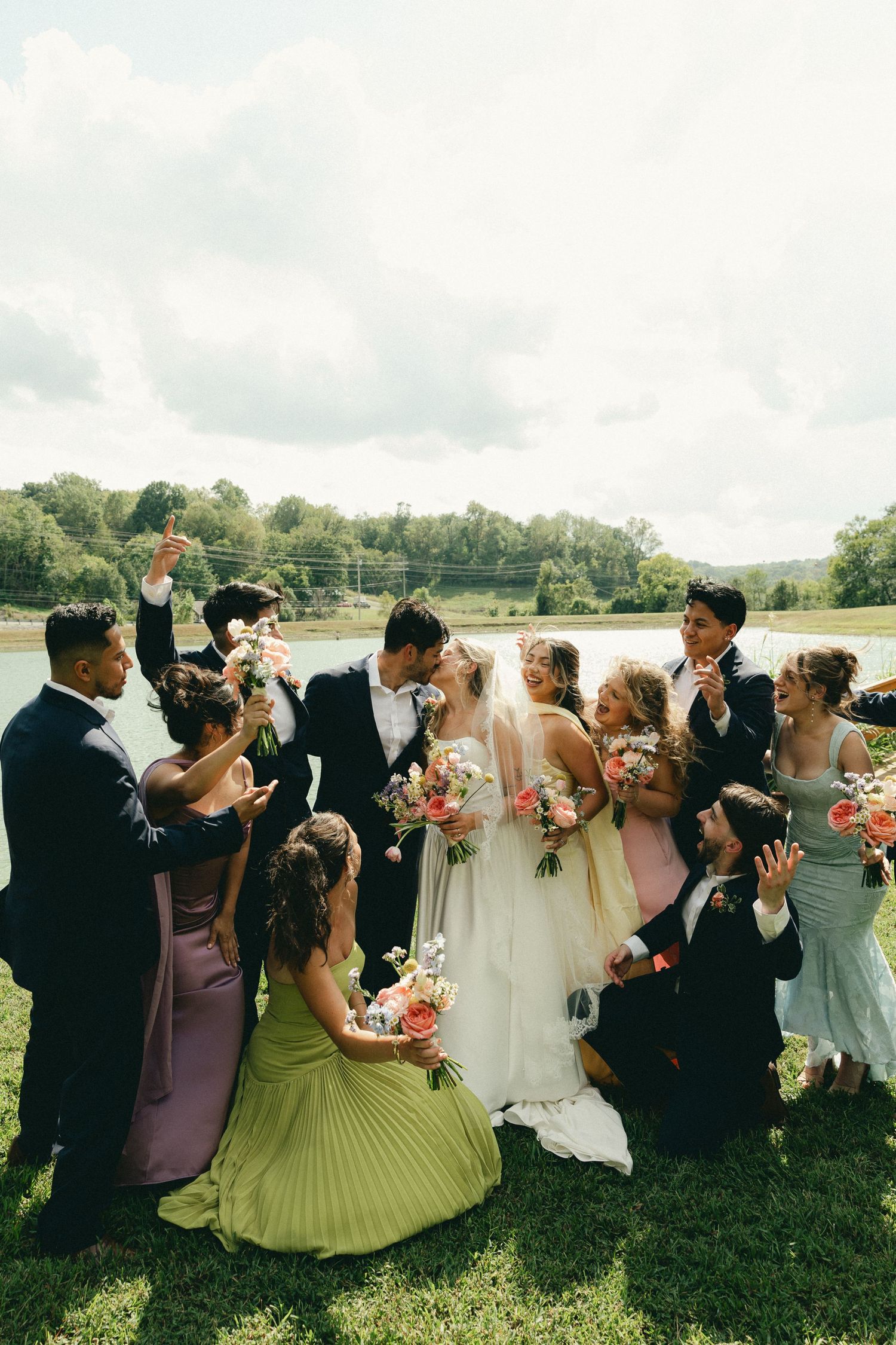 Wedding party celebrating with bride and groom outdoors near a lake, bridesmaids in colorful gowns holding bouquets.
