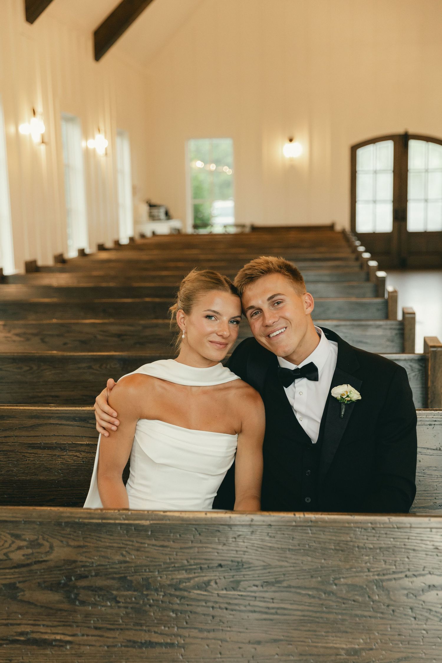 Smiling bride and groom sit together in wooden church pews during their wedding day.