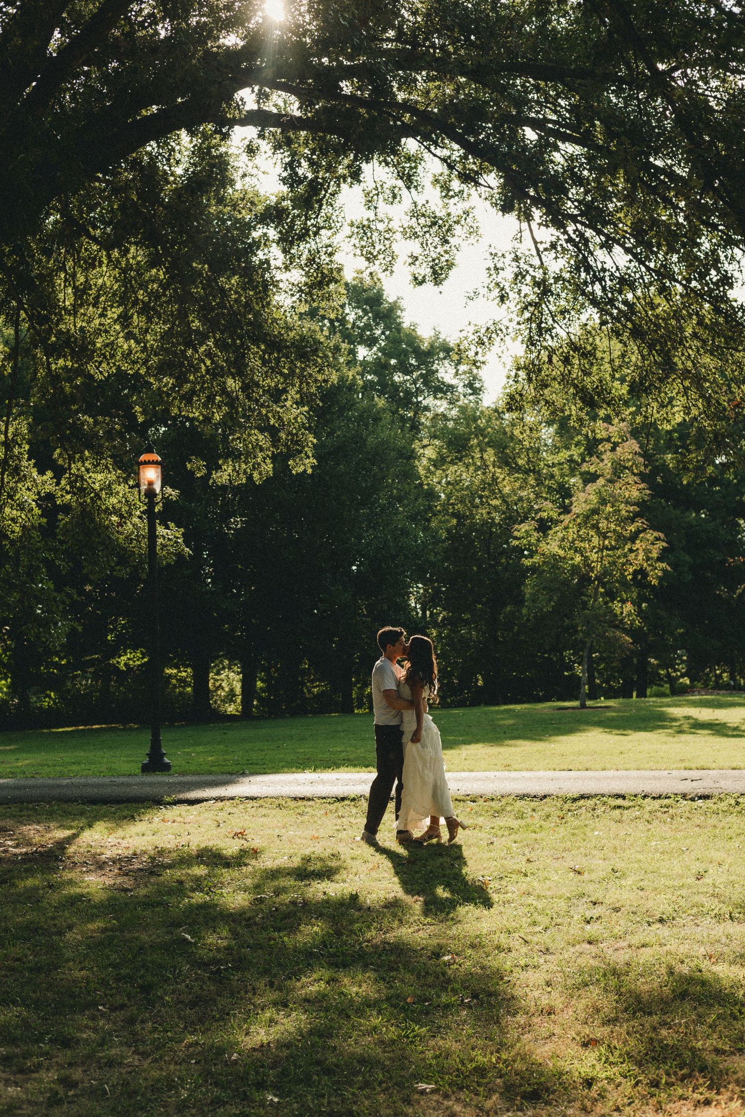 Couple embracing in a sunlit park surrounded by lush green trees and a vintage lamp post on a warm summer day.
