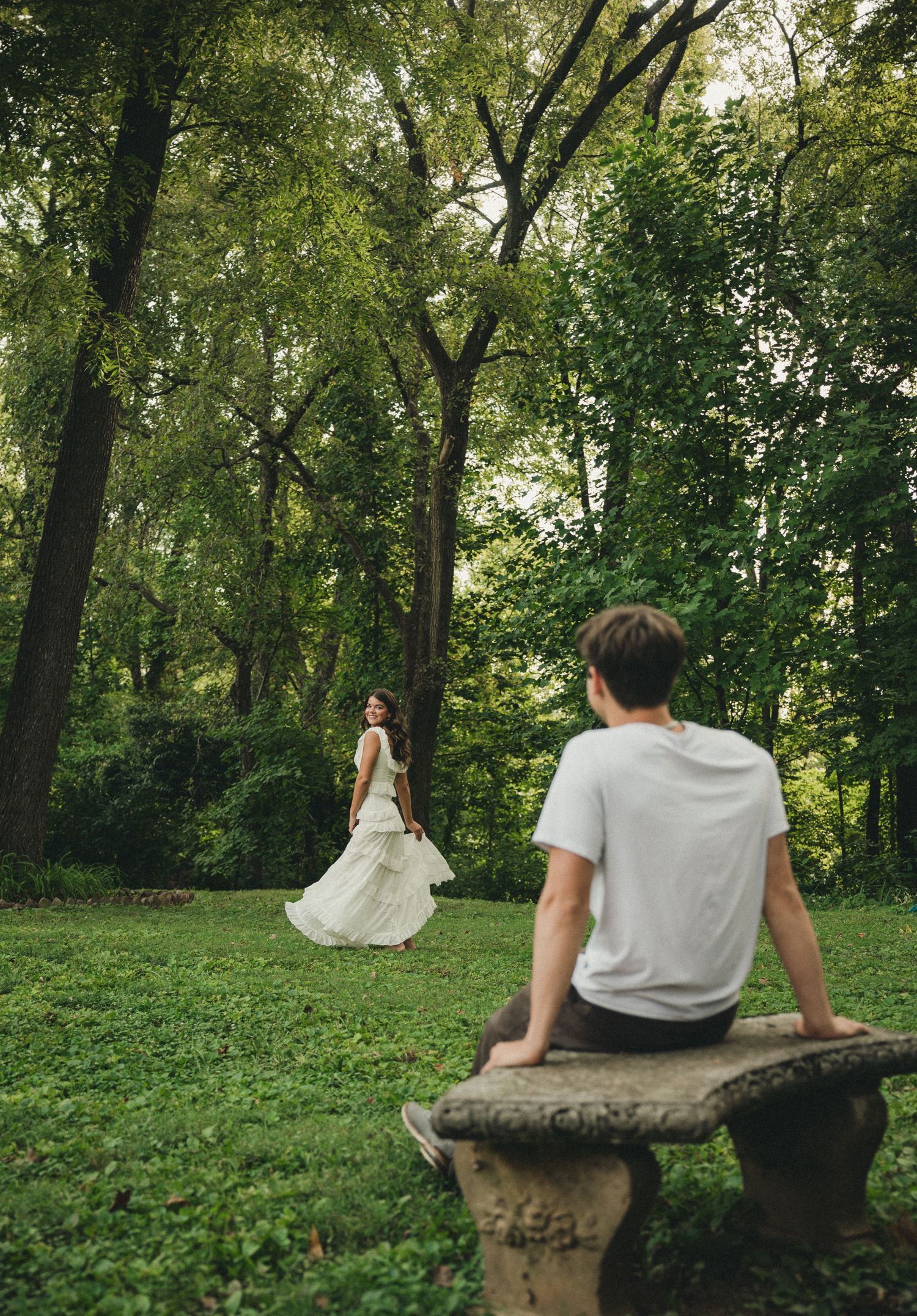 A man sits on a stone bench watching a woman in a white dress twirl in a lush green forest setting.