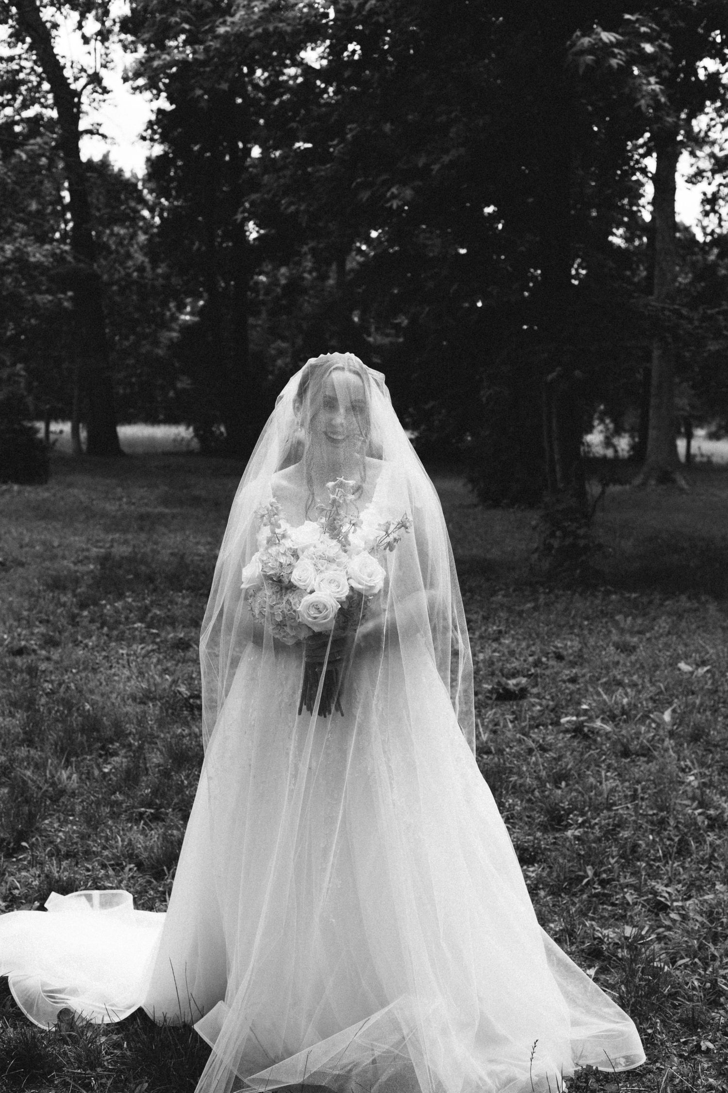 Black and white photo of bride with veil covering face holding bouquet in outdoor park setting.